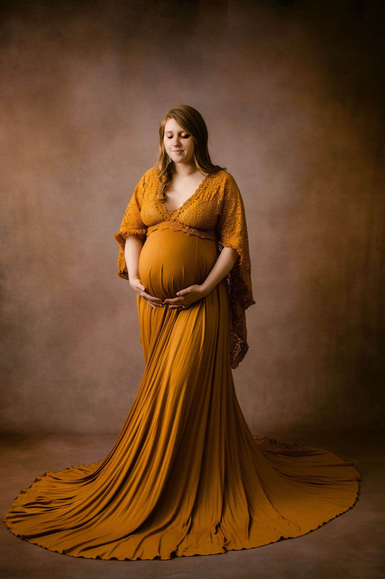 Expectant mother in flowing mustard yellow gown poses gracefully against moody brown studio backdrop for maternity portrait.