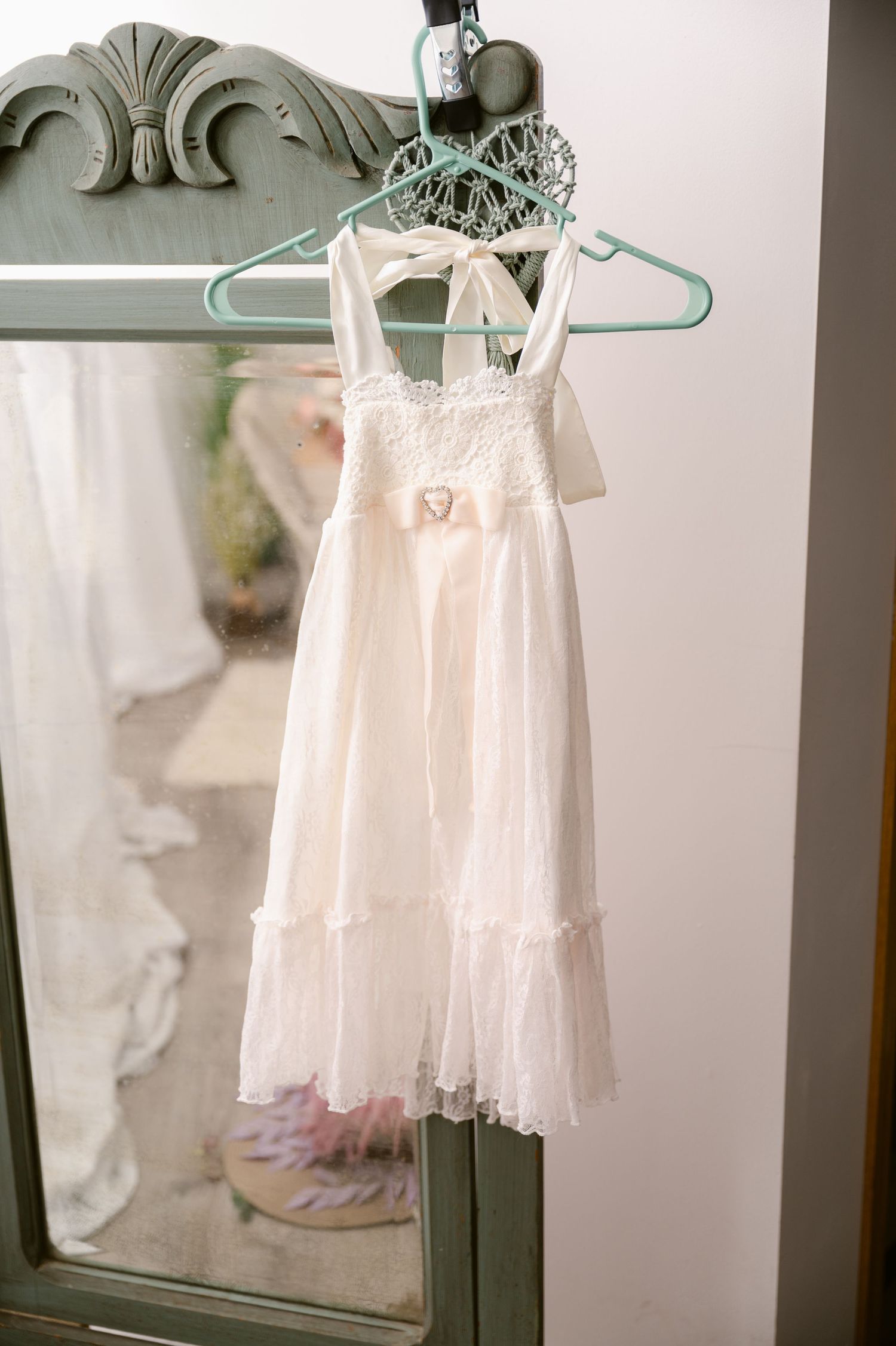 A delicate white lace flower girl dress hangs from a mint green hanger against an ornate vintage mirror.
