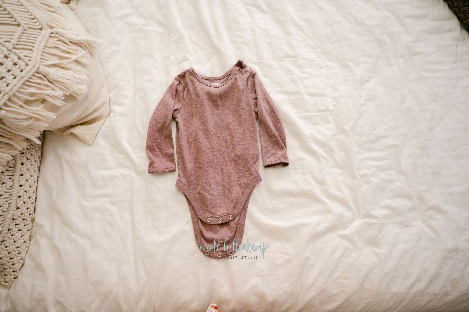 A dusty rose colored long-sleeve baby bodysuit laid flat on white bedding.