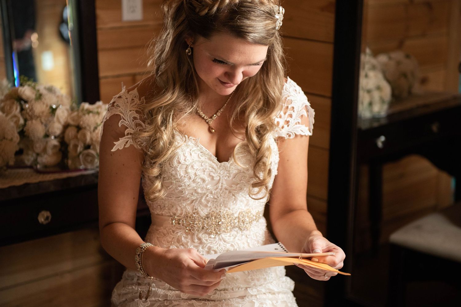 A bride in a lace dress reads a note while sitting in a rustic wooden setting.