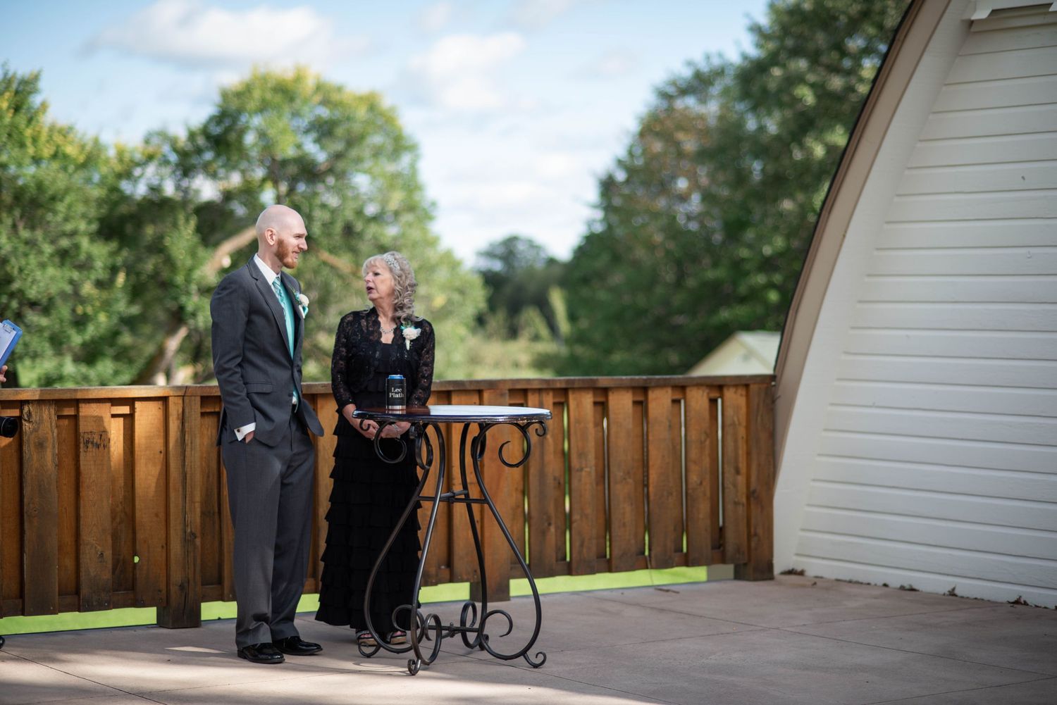 Two people stand on a wooden deck beside a white arched building during an outdoor ceremony.