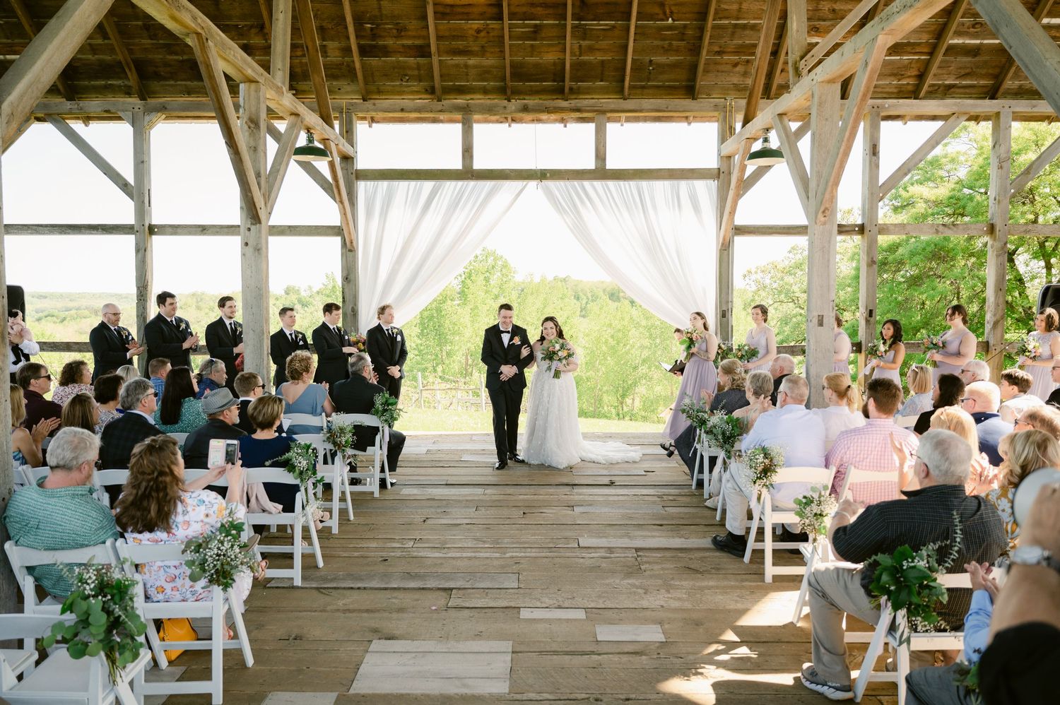 A rustic wooden pavilion filled with guests watching a wedding ceremony with white drapes and floral decorations.
