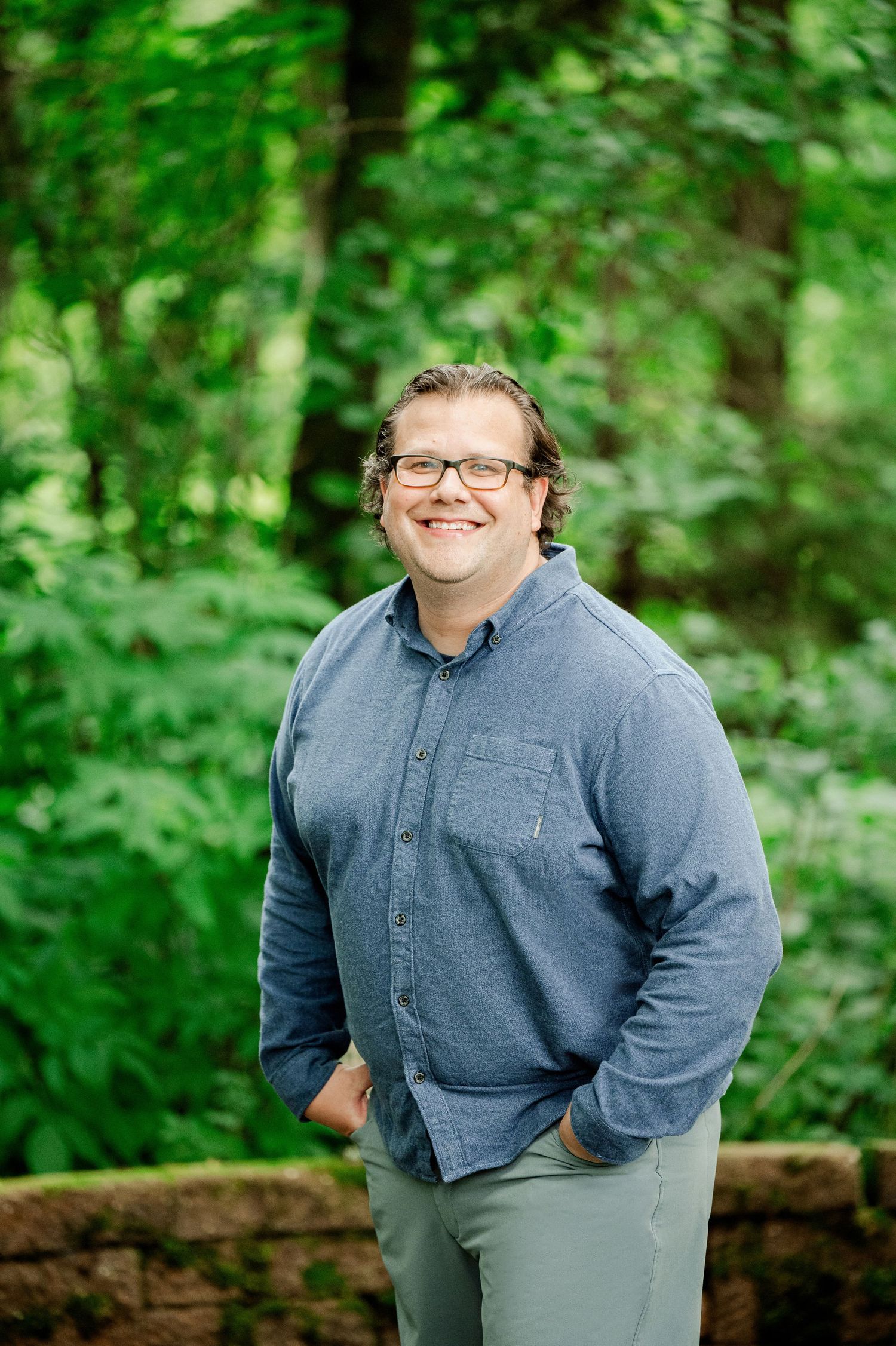 Clear Lake MN Family Photos:Person wearing glasses and a blue button-down shirt smiles warmly while standing in a lush green forest setting.