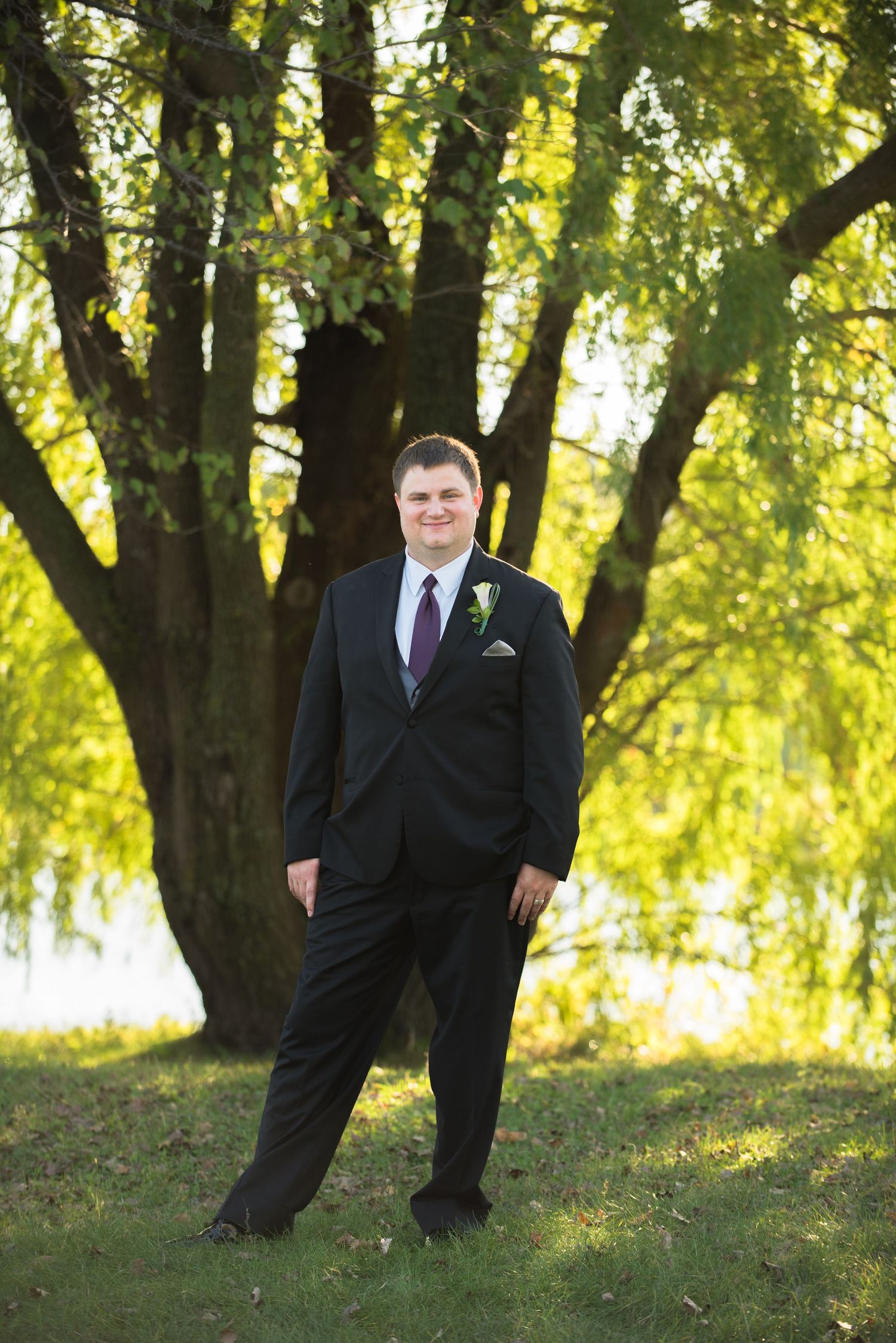 A groom in a black suit poses for a portrait under a large tree with sunlit leaves. Andover wedding at Magestic Oaks Golf Club