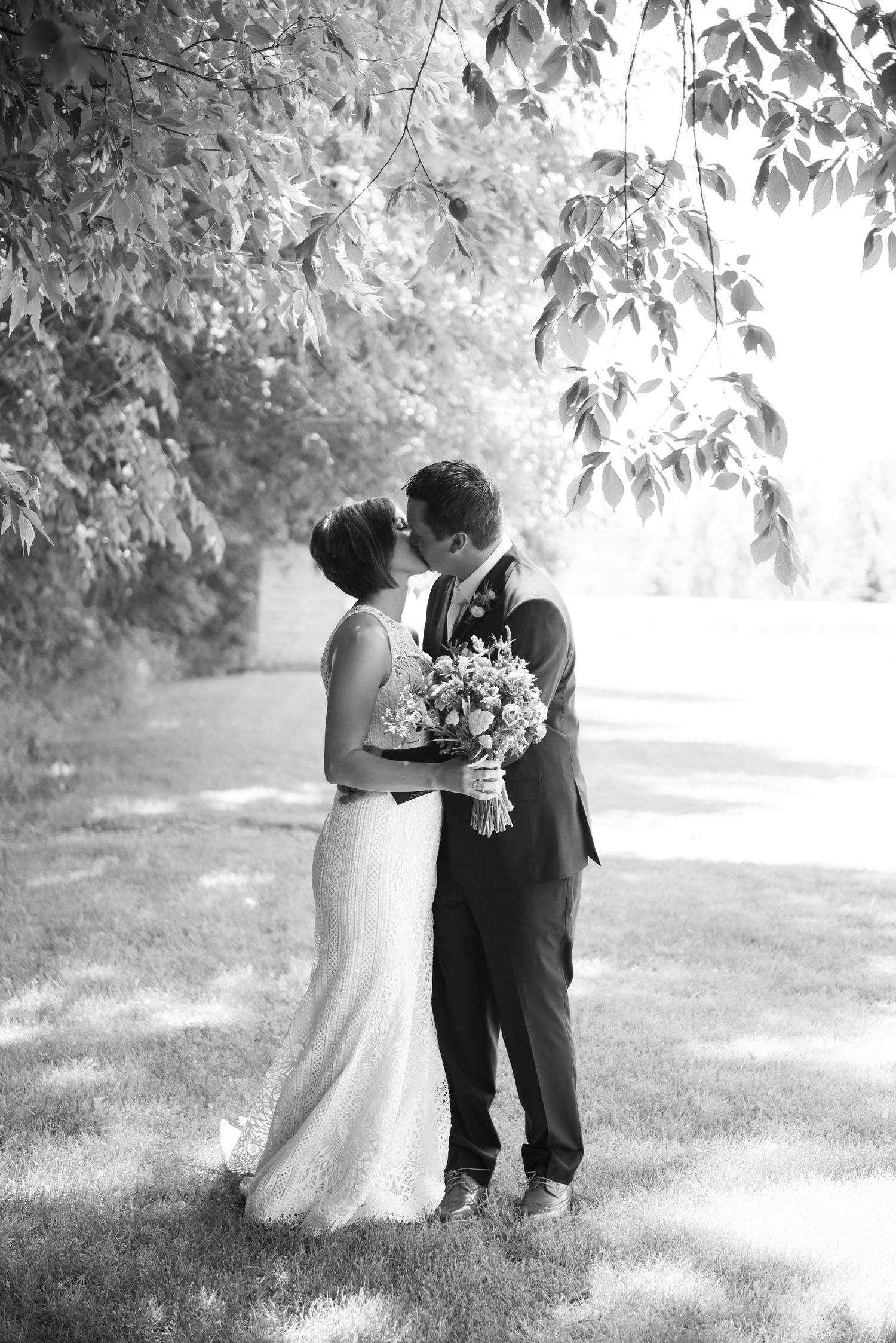 A romantic black and white wedding photo showing a couple embracing under trees while holding a floral bouquet.