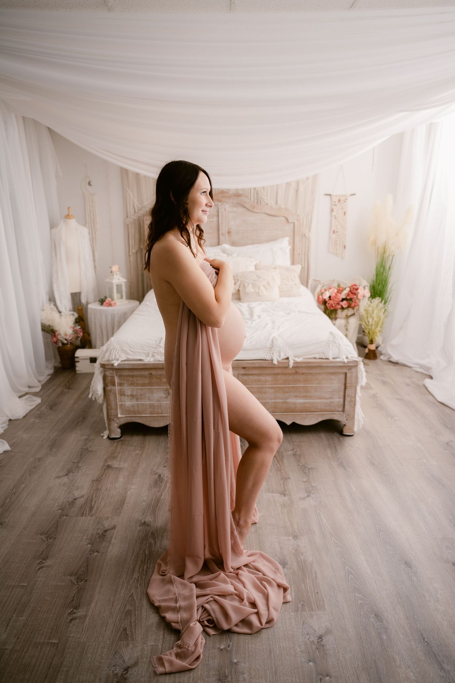 Romantic maternity boudoir photo with pink dress in dreamy white bedroom setting with vintage bed frame.