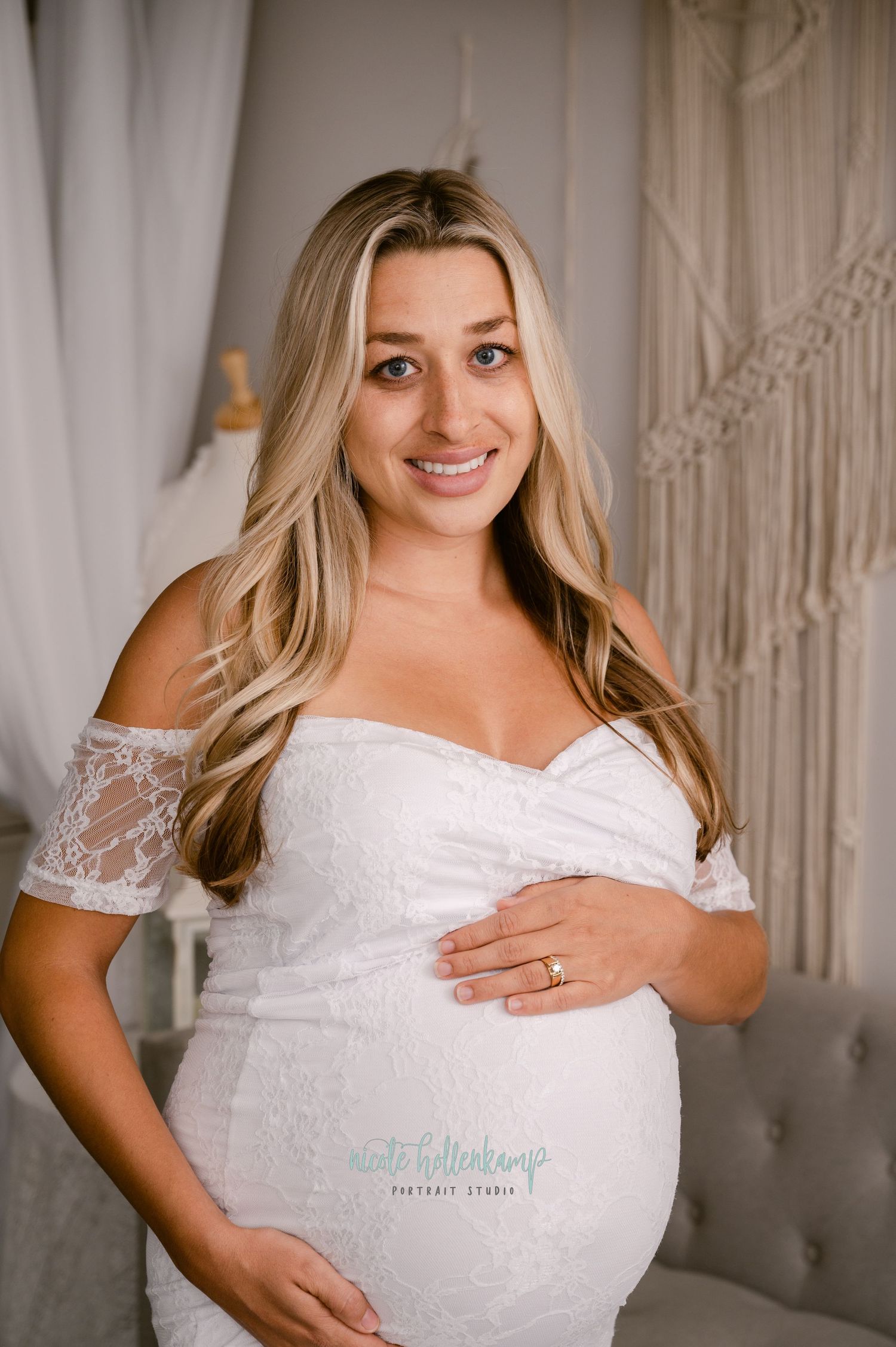 Expectant woman wearing white lace off-shoulder maternity dress poses against macrame backdrop in indoor photo session.