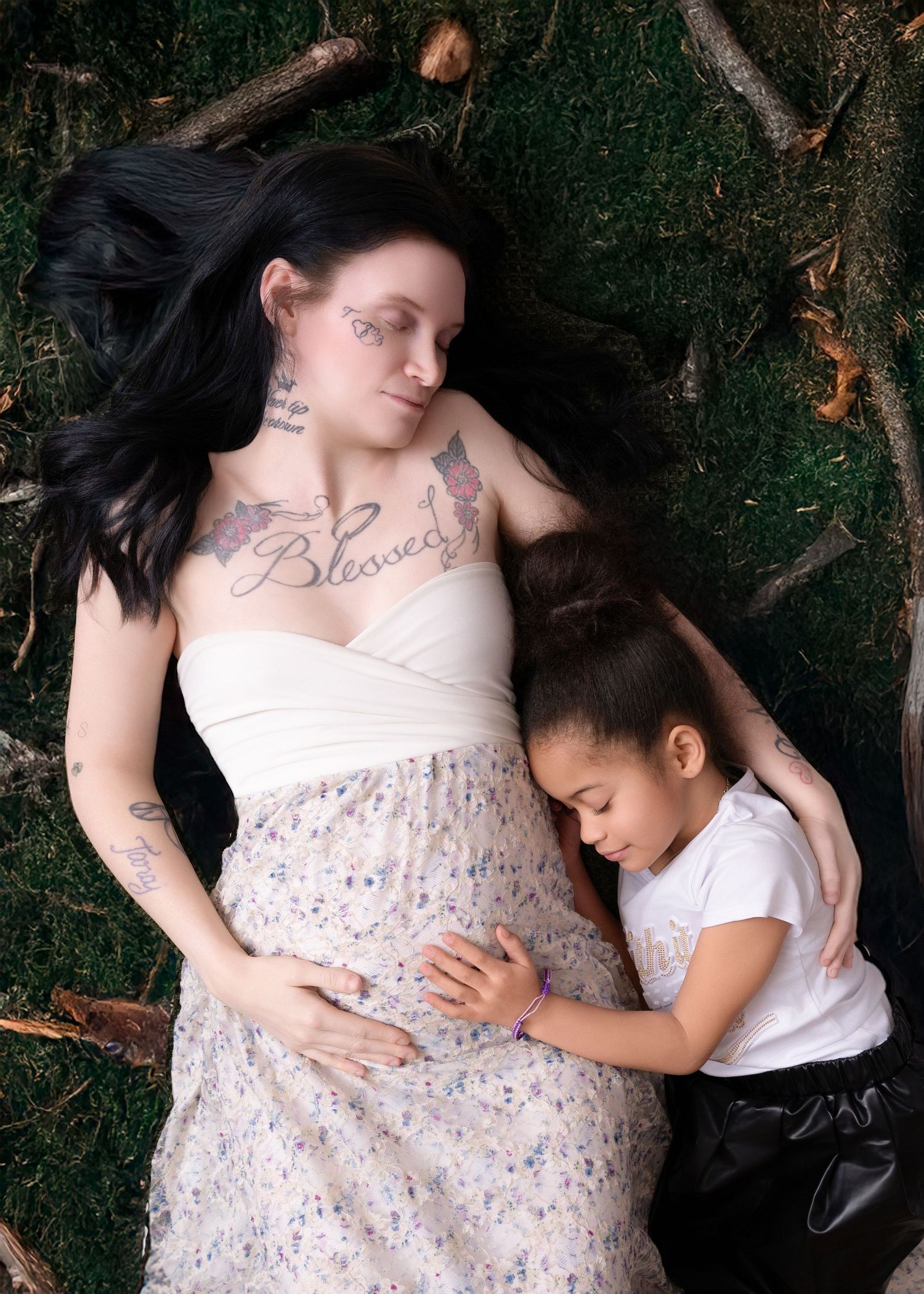 Mother in white dress rests on grass while child embraces her pregnant belly.
