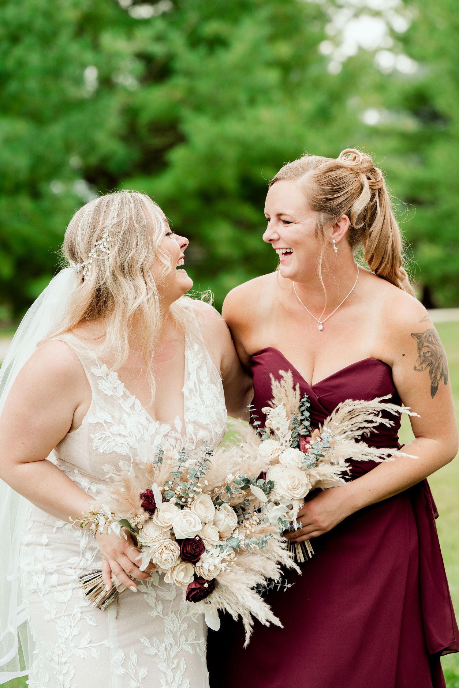 A bride in white lace and bridesmaid in burgundy share a joyful laugh while holding a neutral-toned floral bouquet outdoors.