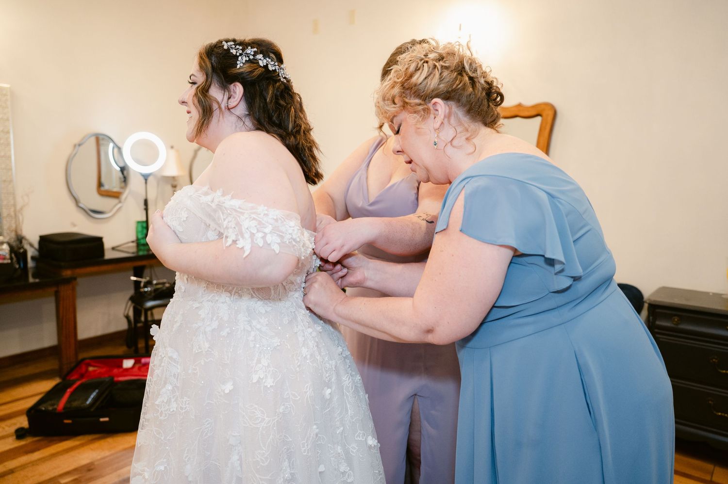 Wedding attendants help with final dress adjustments in a preparation room.