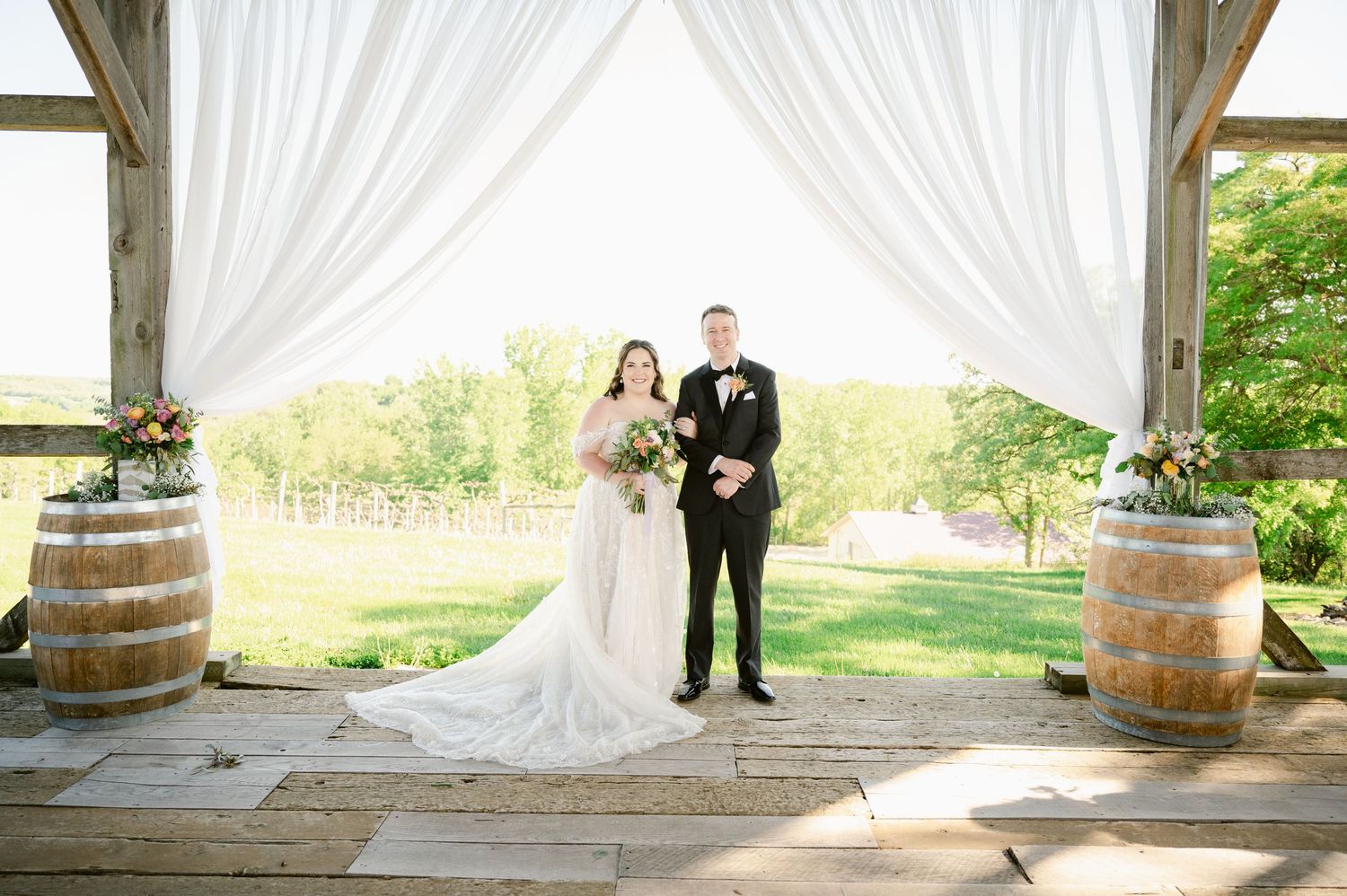 Newlyweds standing in a rustic wooden pavilion decorated with white drapes and wine barrels.
