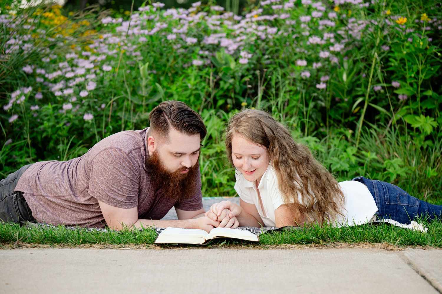 Couple lying on blanket reading together surrounded by blooming wildflowers on concrete path.