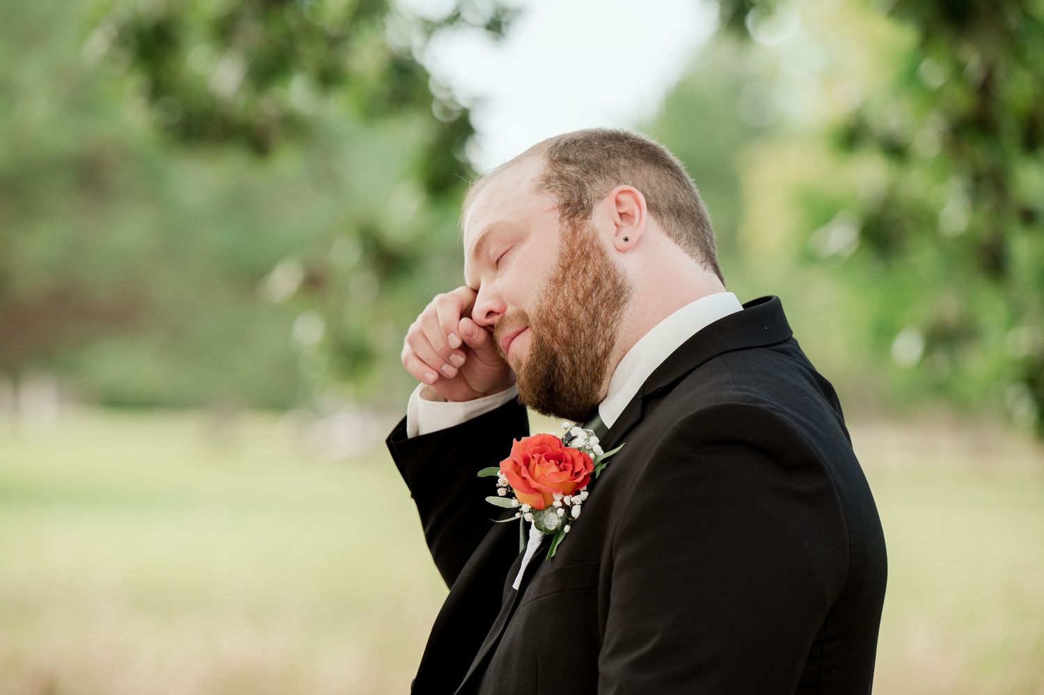 The Grands at Mulligans- Wedding Photos: Groom wearing a traditional black tuxedo and coral flower stands under green foliage outdoors.