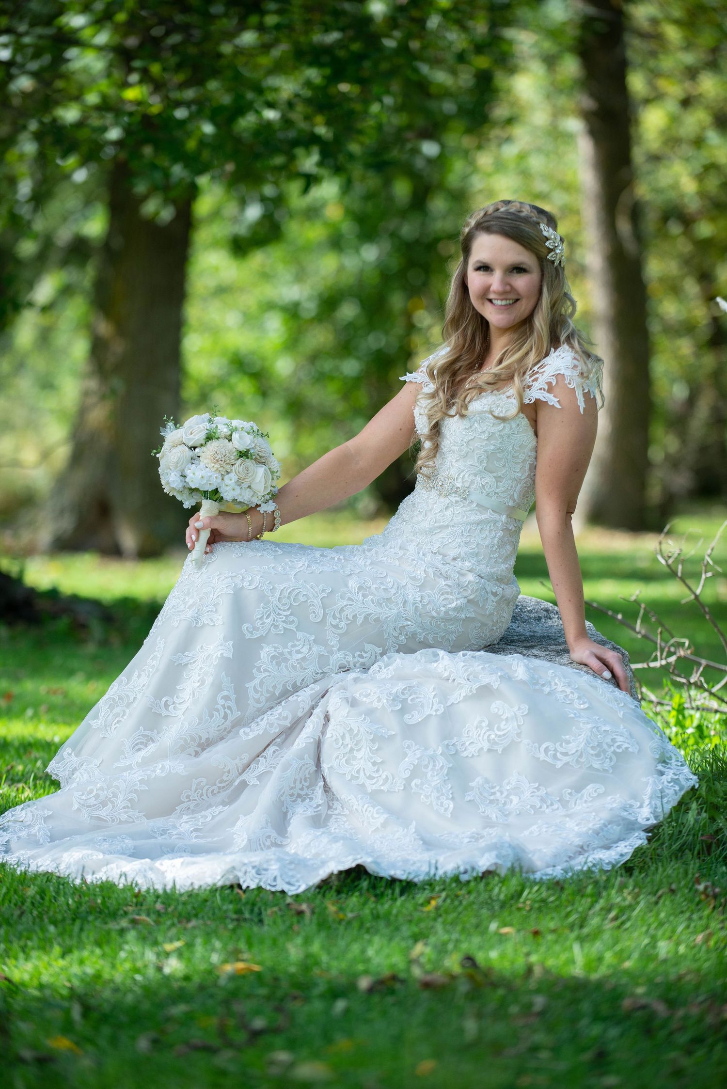 A bride in a detailed lace wedding dress sits gracefully on grass in a garden setting with a white floral bouquet. Rum River Barn Milaca Min