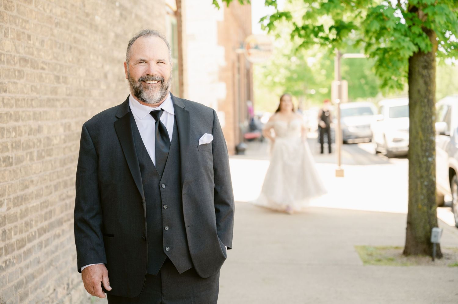 A groom in a black tuxedo poses against a brick wall while waiting for the first look.
