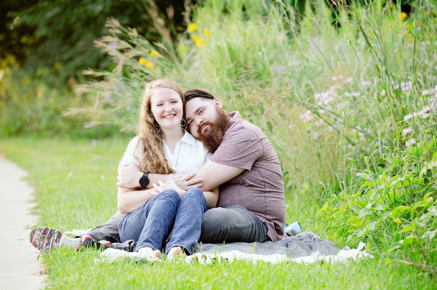 St cloud area engagement photos. Two people sitting together on a blanket outdoors with tall grass and greenery in the background.