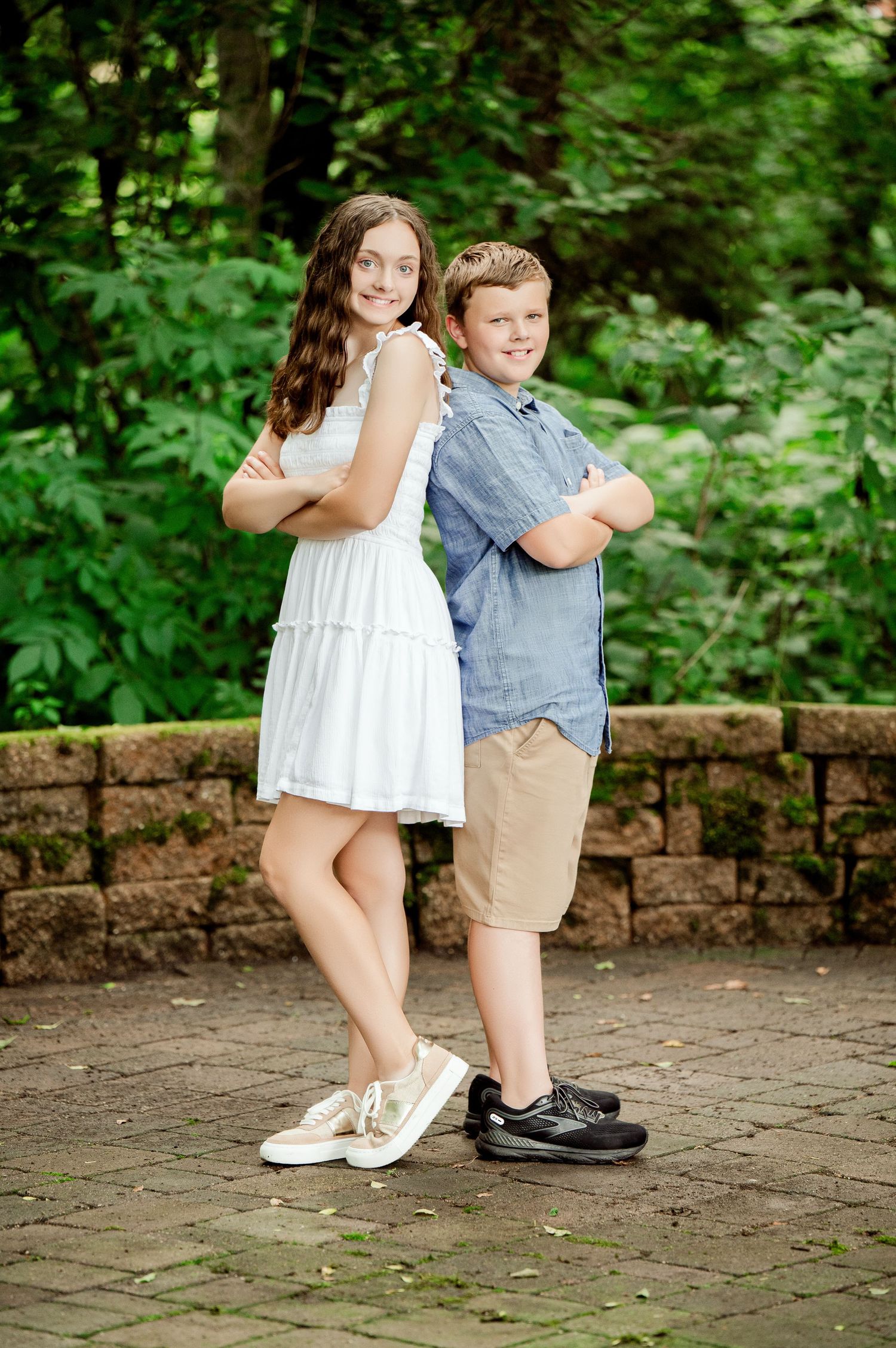 Two people pose back-to-back on a brick pathway surrounded by green foliage in white dress and casual attire.