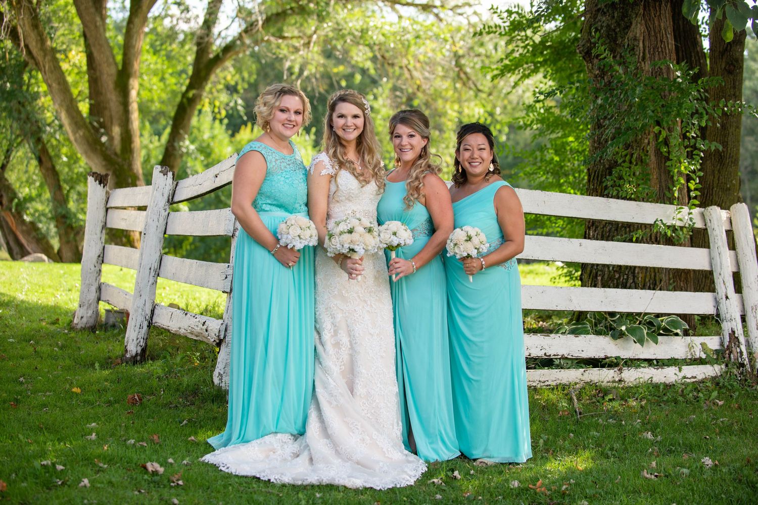A bridal party in turquoise dresses poses by a white fence in a garden setting. Rum River Barn Milaca Minnesota Weddings