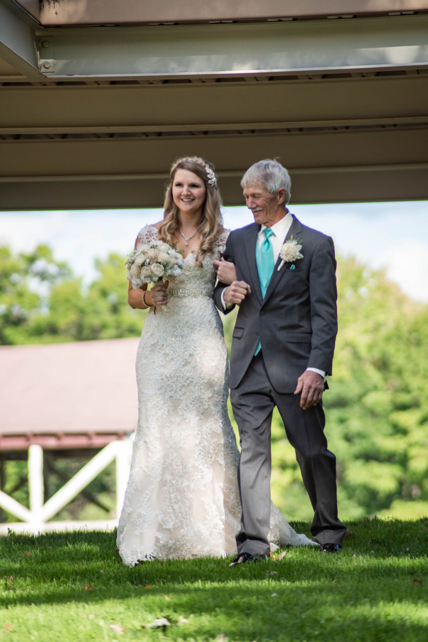 A wedding couple in formal attire poses outdoors on a grassy hill beneath a wooden structure.
