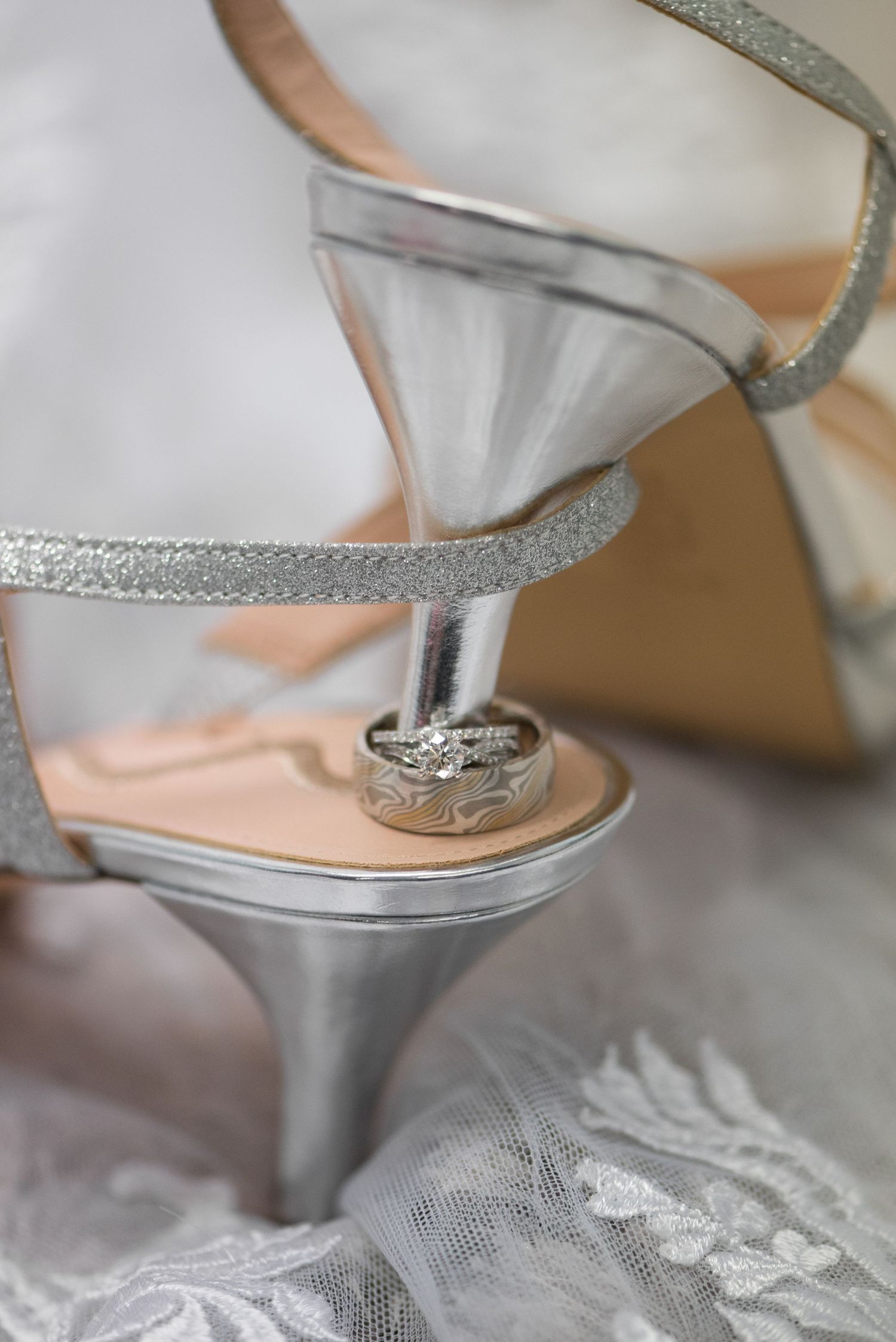 Silver glitter wedding shoes with diamond rings balanced on the heel against lace fabric. Andover wedding at Magestic Oaks Golf Club