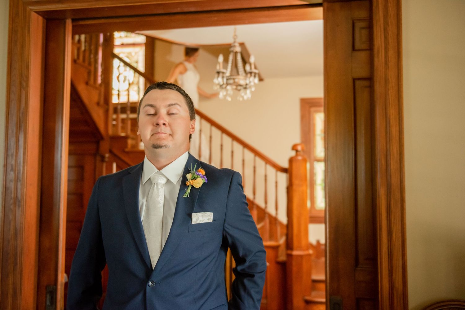 A groom in a navy blue suit and pale tie stands in a wooden doorway with stairs and chandelier visible behind him.