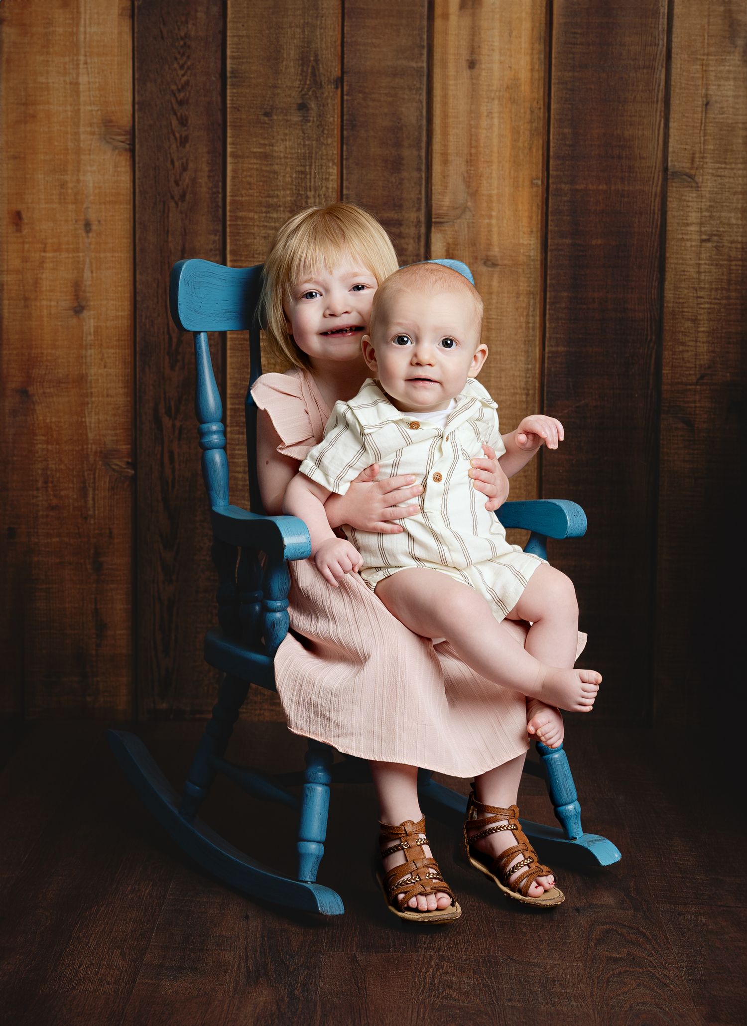 Two children sitting together on a blue wooden rocking chair against rustic wood paneling.