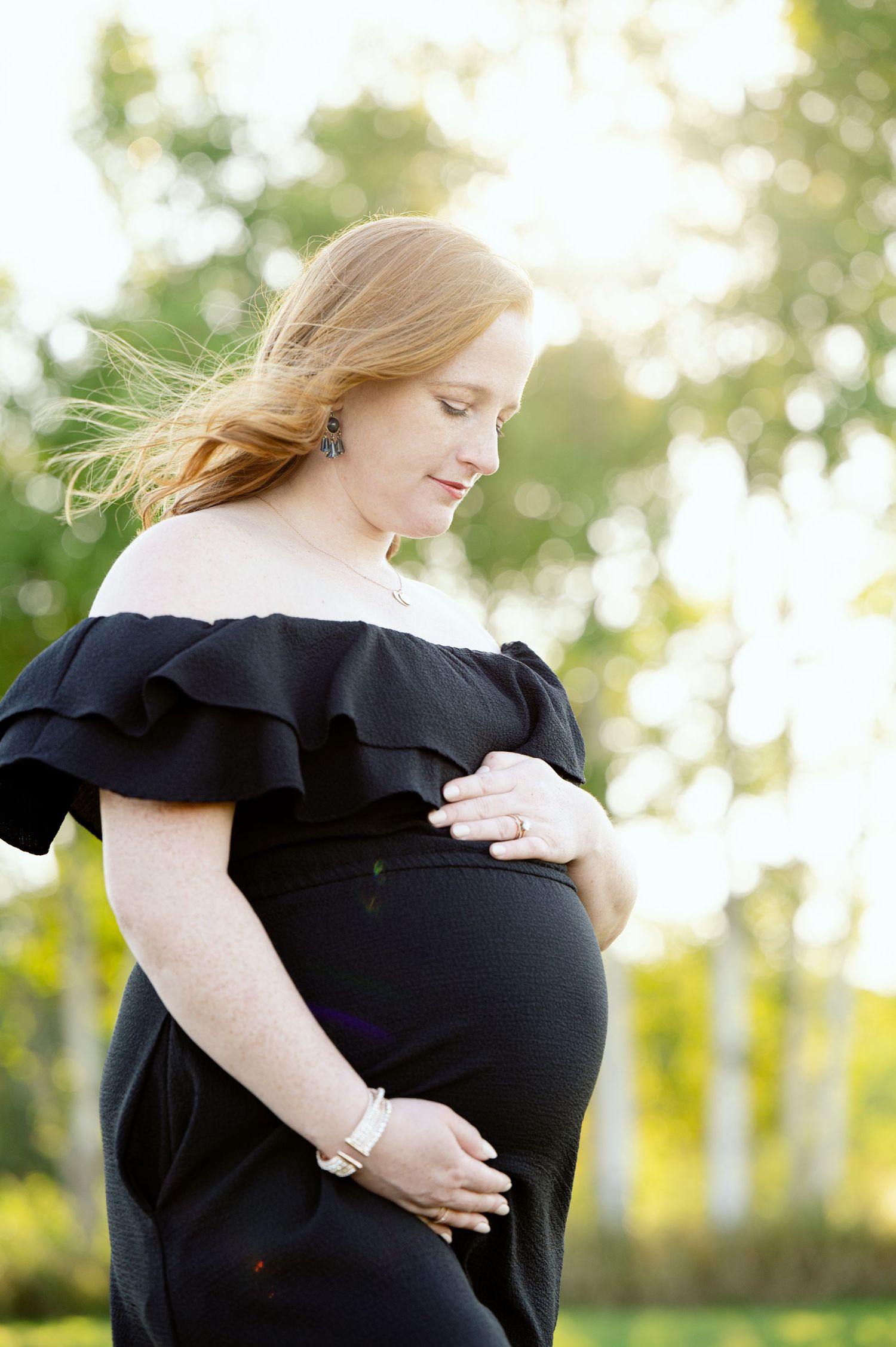 A side profile of an expectant mother in black dress with ruffled shoulders amid golden sunlight.