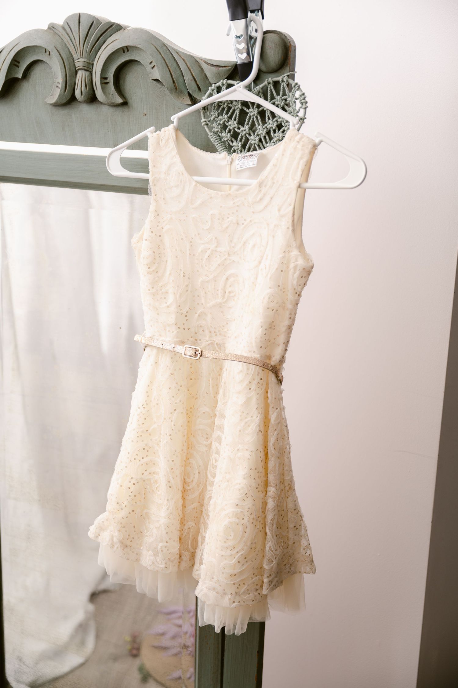 A cream-colored lace party dress with a fitted bodice and flared skirt hangs on a white plastic hanger against a light wall.