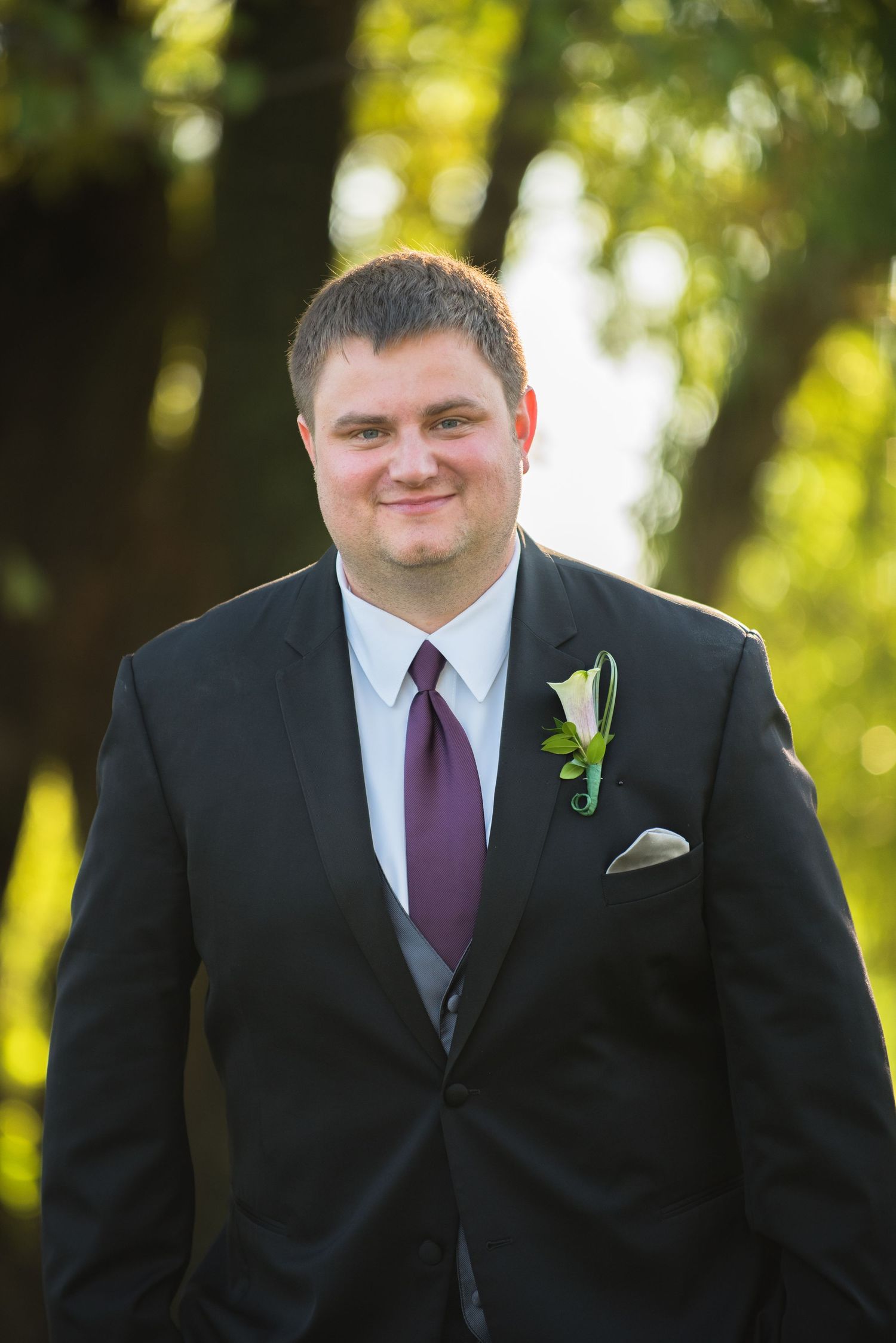 A groom wearing a black suit with purple tie and white boutonniere smiles at the camera. Andover wedding at Magestic Oaks Golf Club