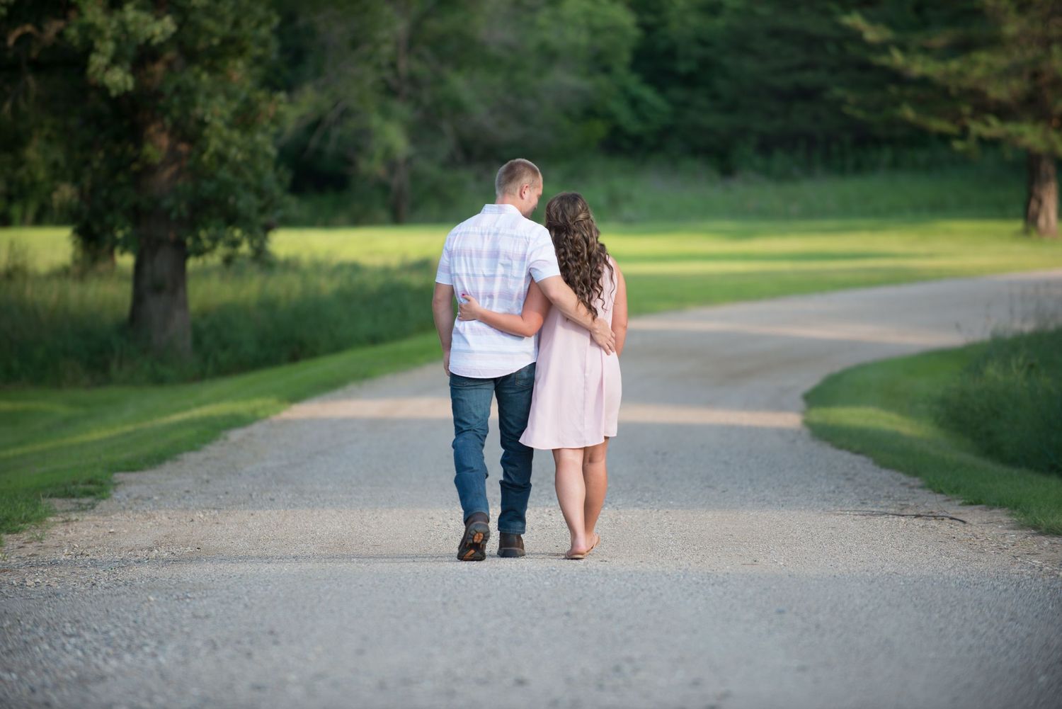 A couple walks hand in hand down a winding gravel path surrounded by green trees and grass.