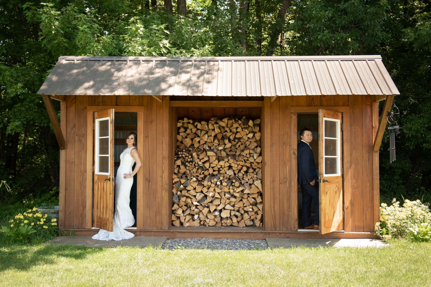 A rustic wooden storage shed with stacked firewood and a door sits nestled among green trees in a backyard setting.