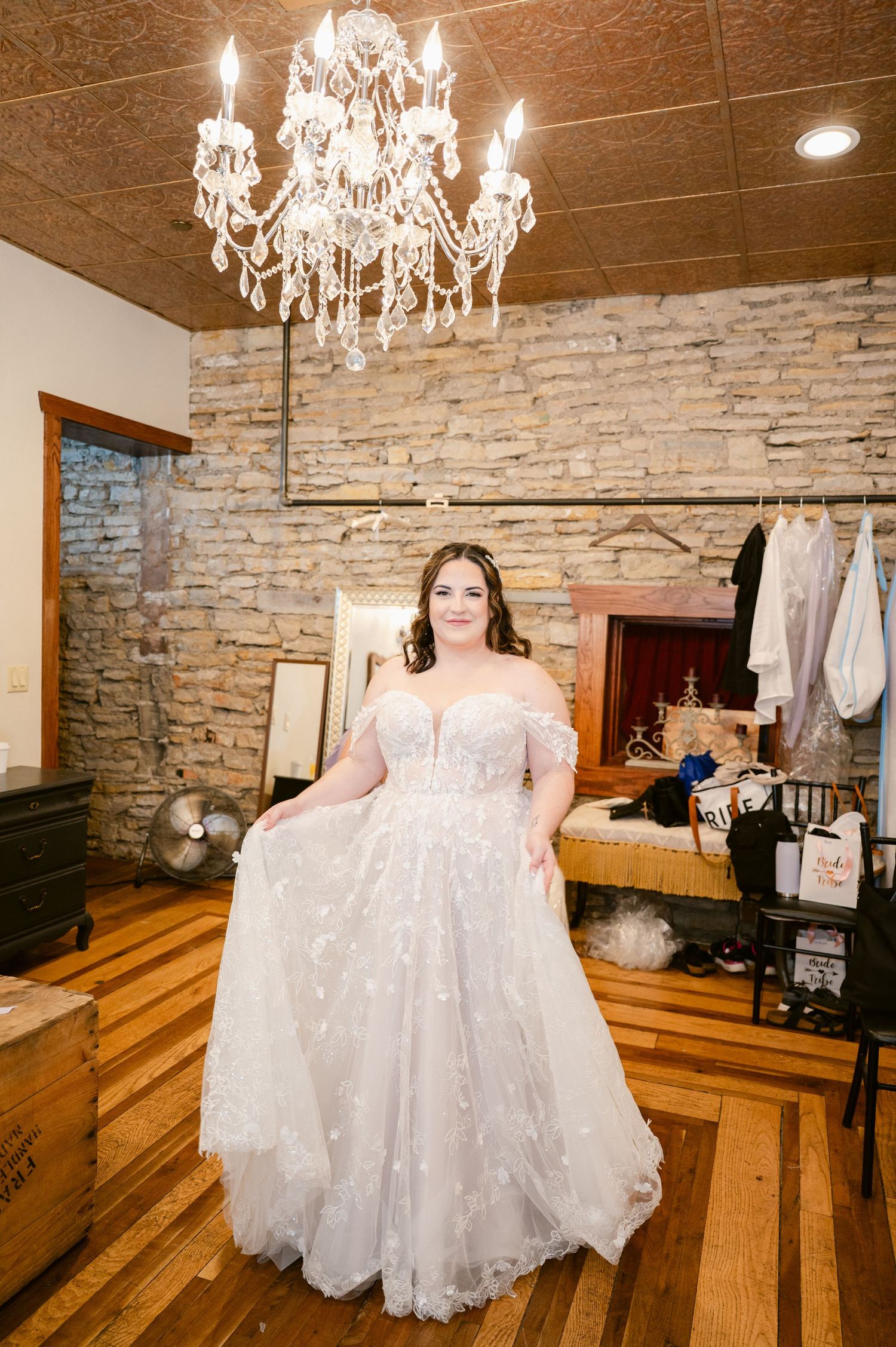 Beautiful off-shoulder lace wedding gown shown in dressing room with elegant crystal chandelier and stone wall backdrop.