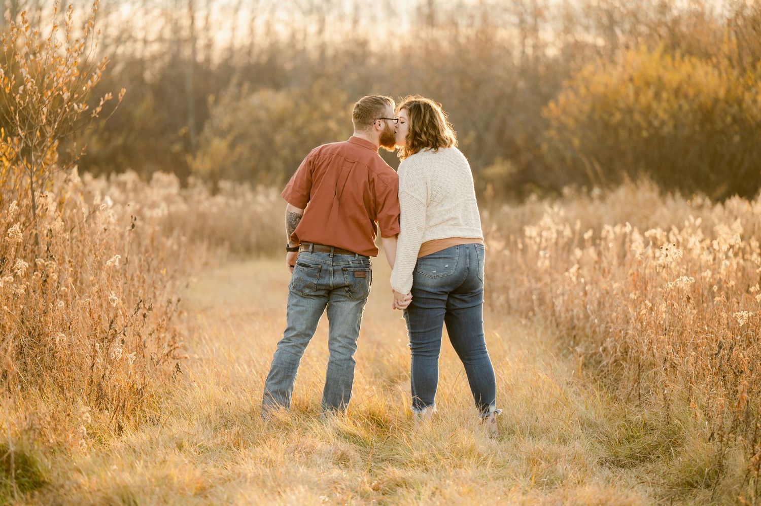 Two figures in casual attire walk through a golden field at sunset holding hands as warm light illuminates the landscape.