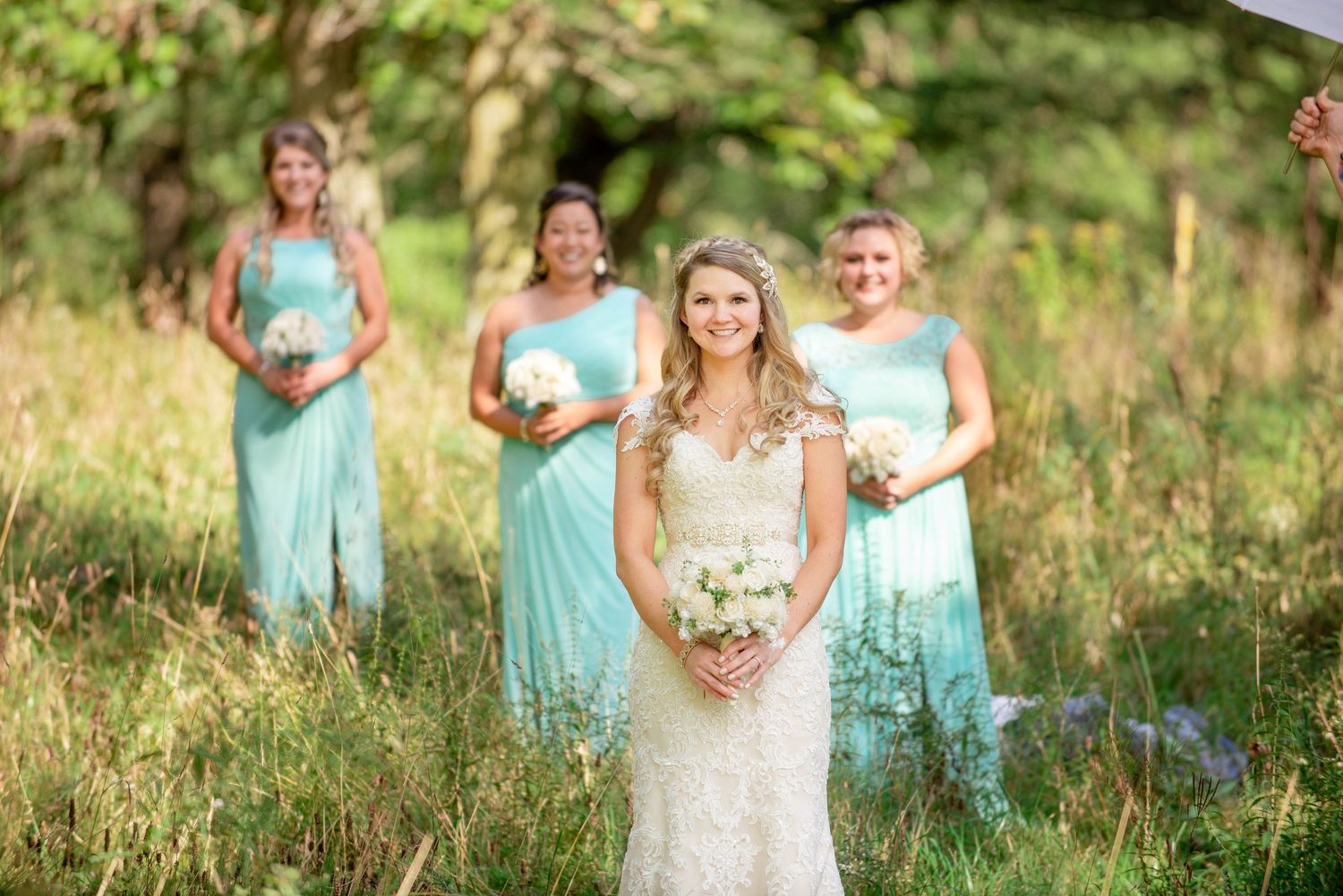 Bride in lace gown stands in field with bridesmaids wearing turquoise dresses. Rum River Barn Milaca Minnesota Weddings