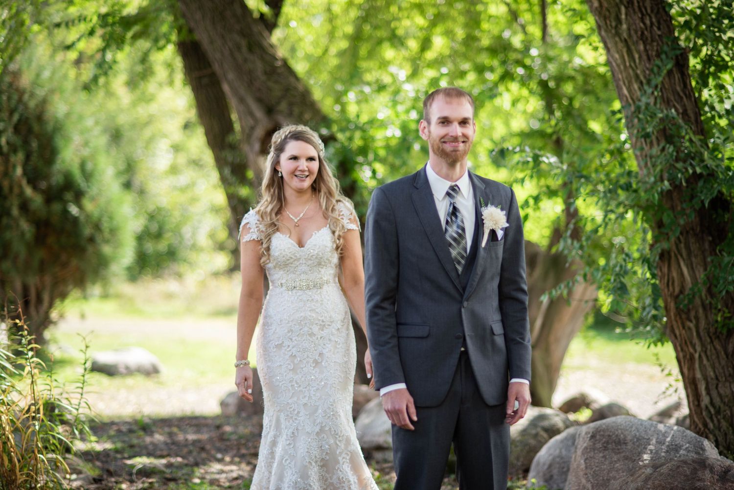 A couple in wedding attire walks and smiles together in a lush green outdoor setting during their celebration.
