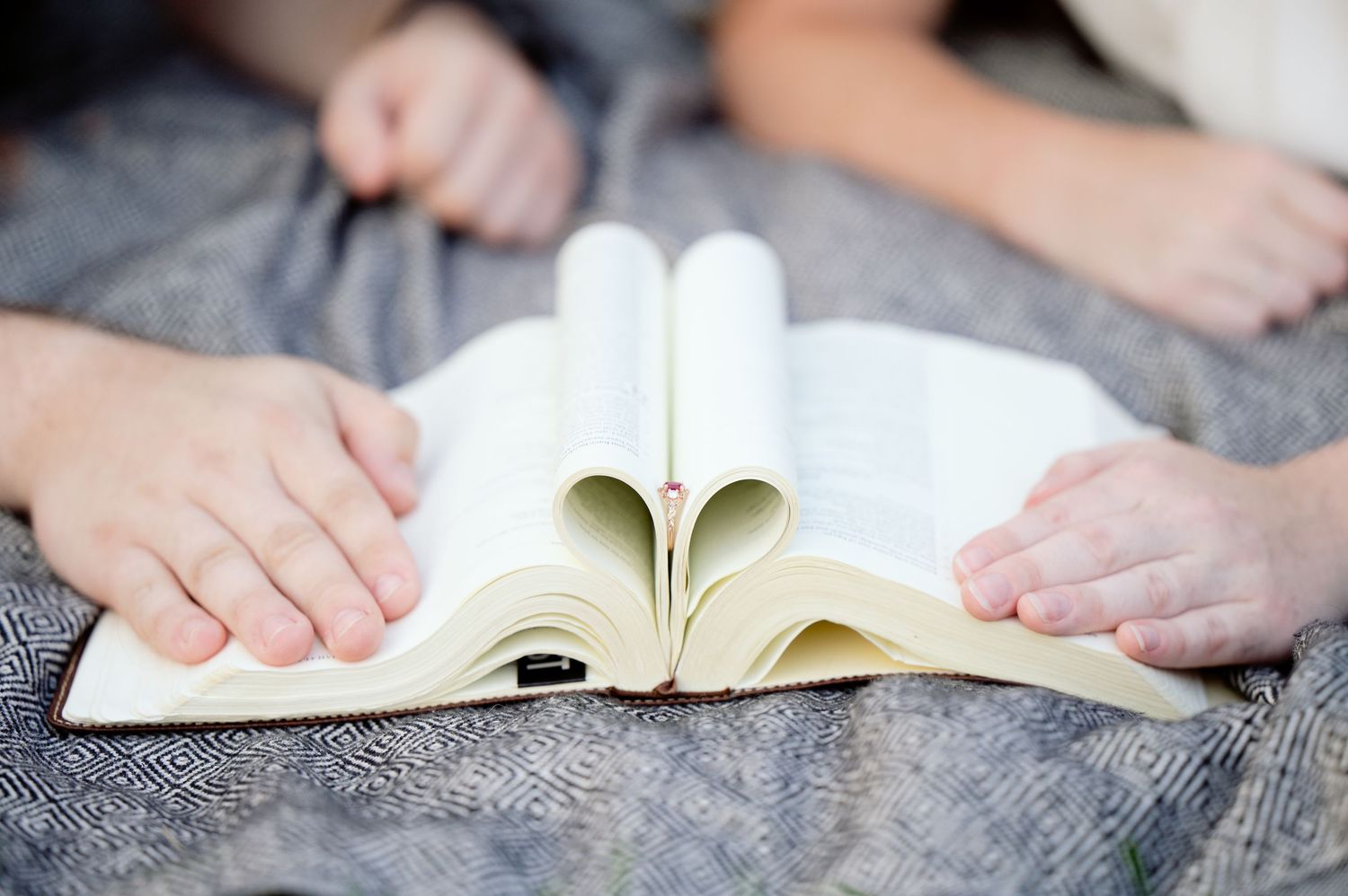 Close-up of hands holding open book pages folded into heart shape on gray textile surface.