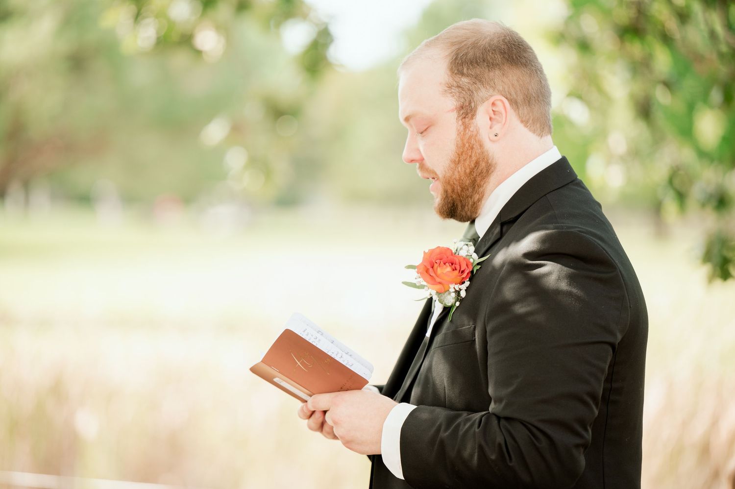 The Grands at Mulligans- Wedding Photos: A bearded groom in a black suit reads personalized vows at an outdoor