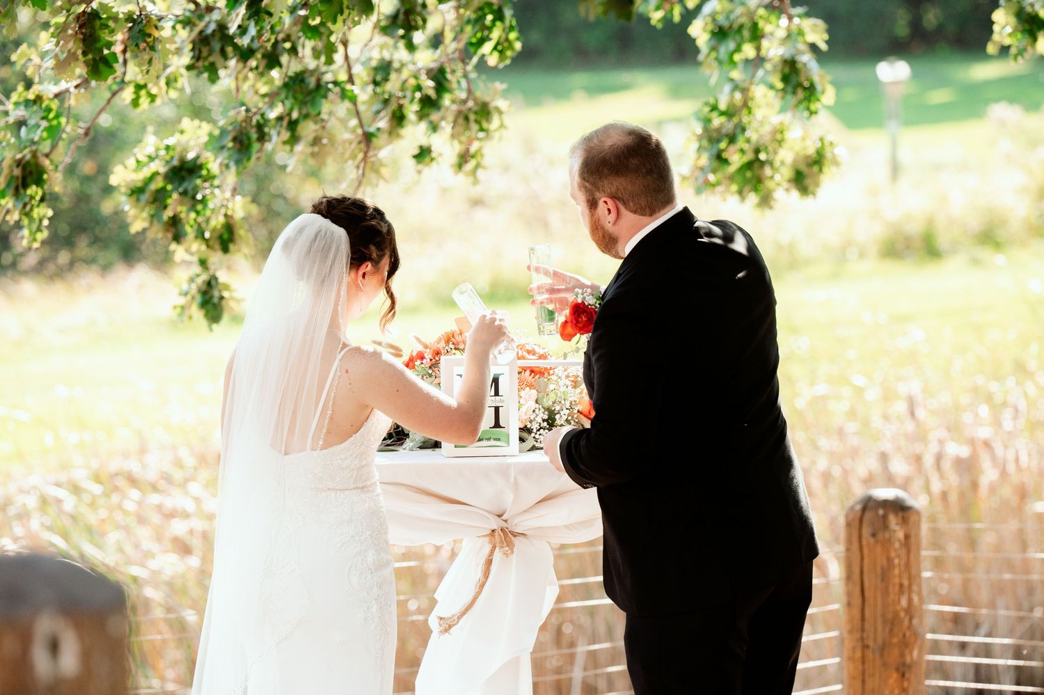 A couple in wedding attire sign documents together under flowering tree branches on a sunny day.