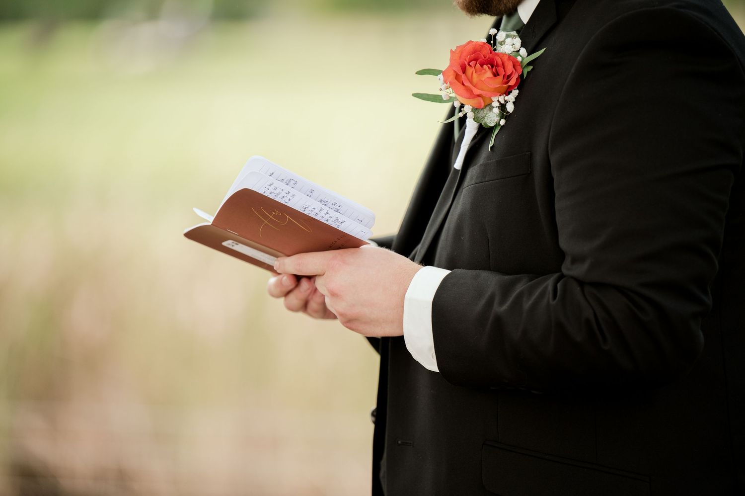 The Grands at Mulligans- Wedding Photos: in black suit holds ceremony book while wearing a coral rose boutonniere during outdoor ceremony.