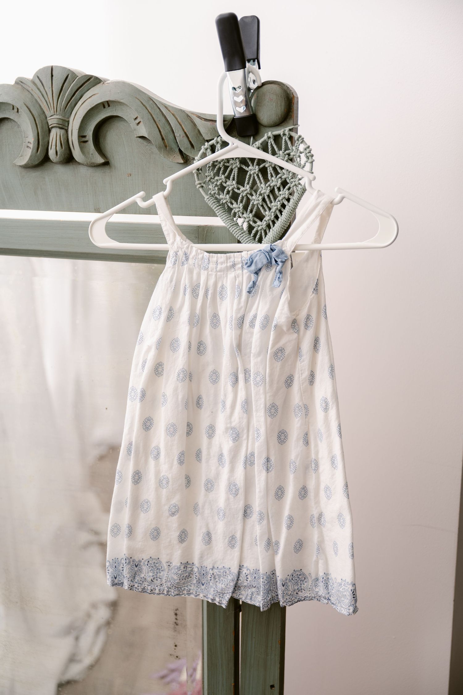 White lace-trimmed sundress with delicate blue floral pattern hangs on white plastic hanger against gray wall.
