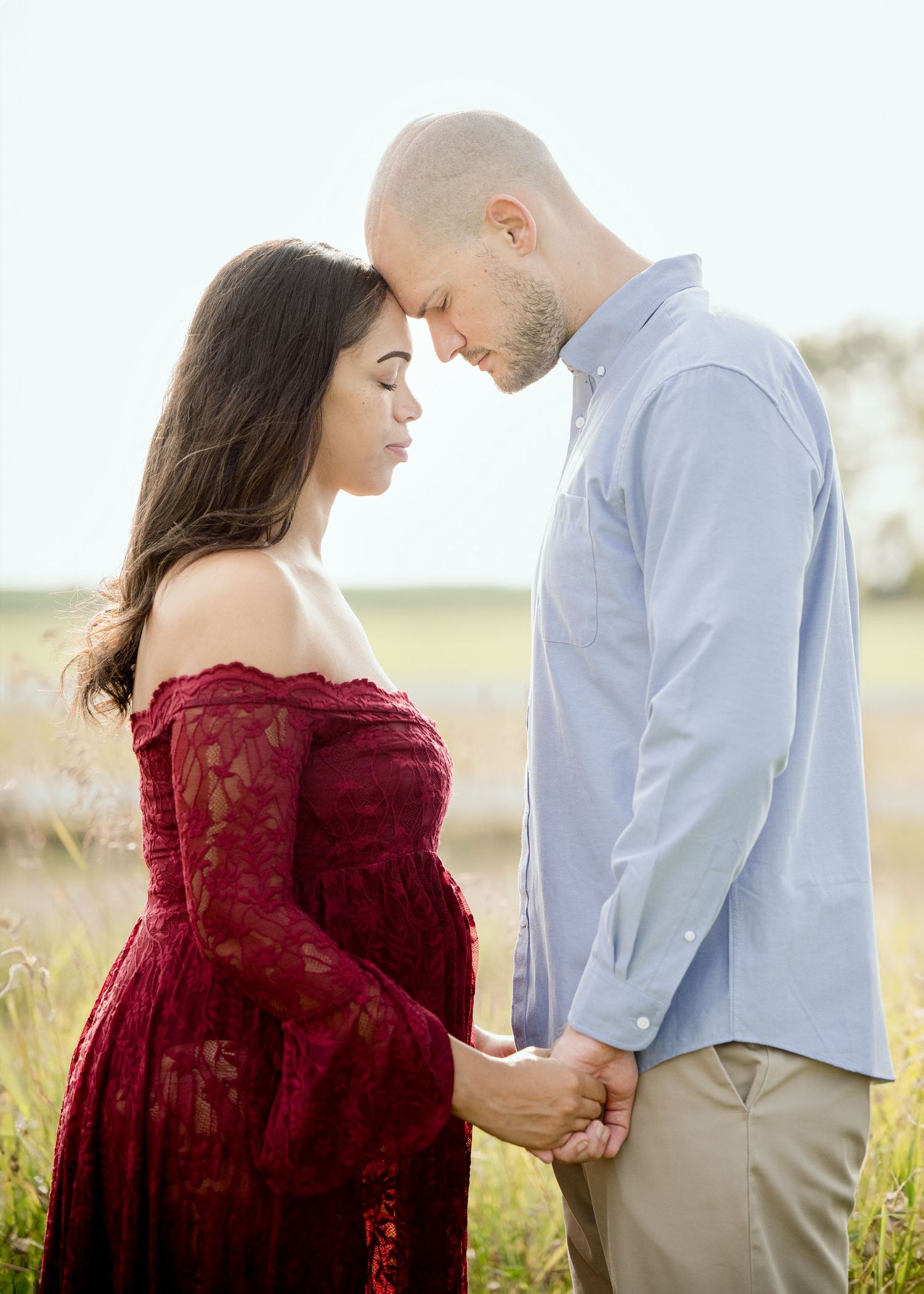 Two people share an intimate moment in a field, wearing a burgundy lace dress and light blue shirt.