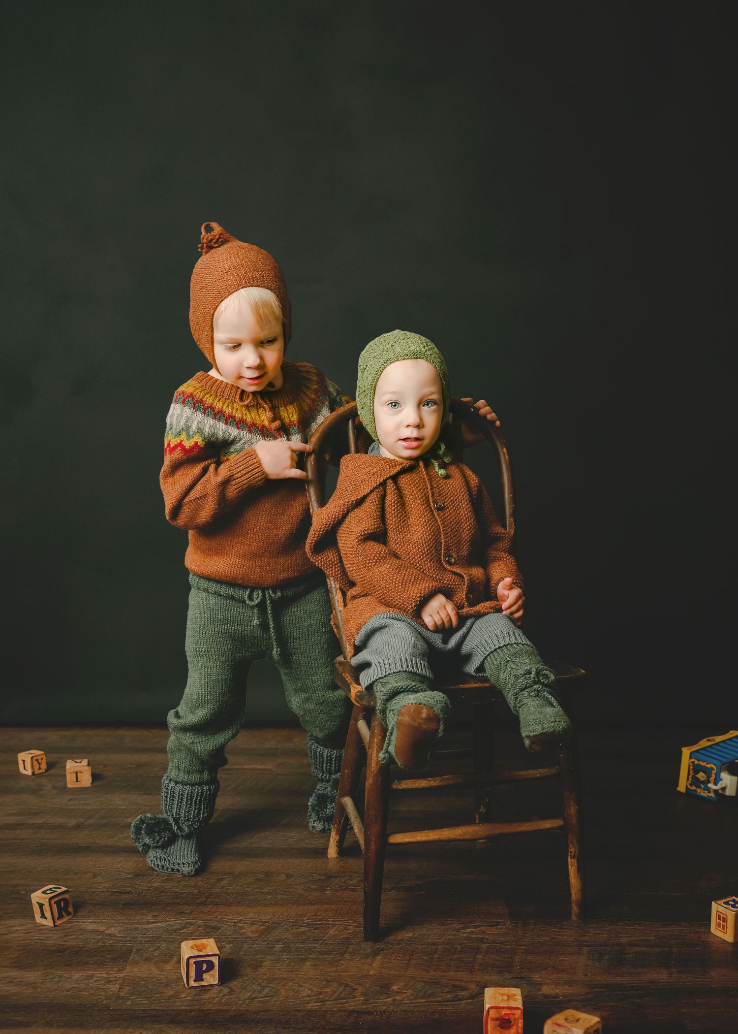 Two children in brown sweaters pose with wooden chair and alphabet blocks.