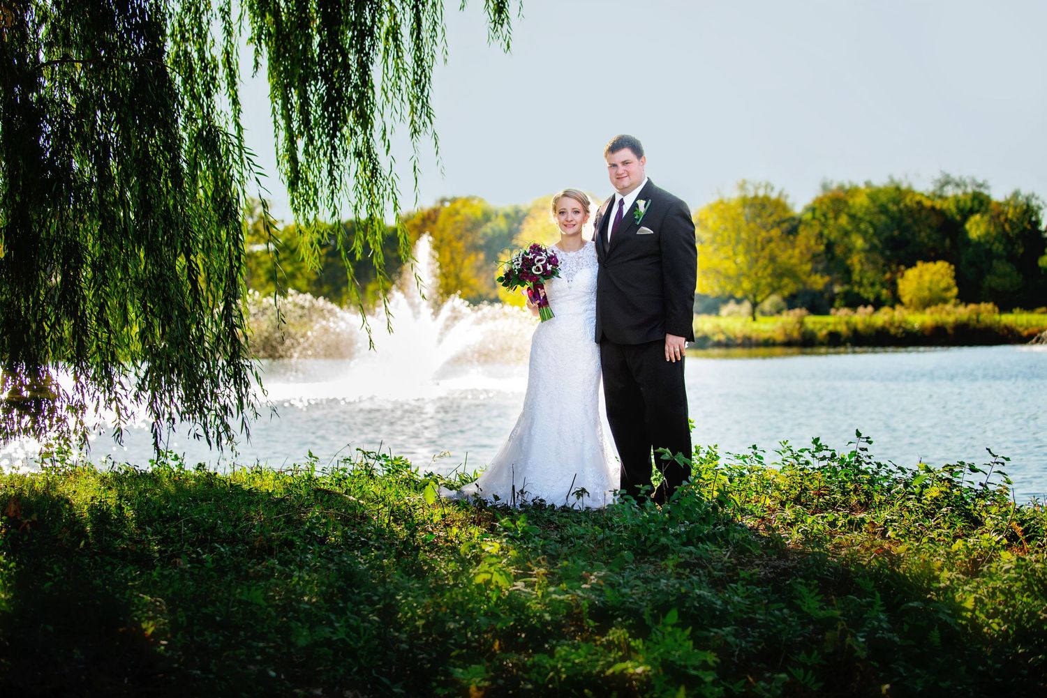Panoramic view of newlyweds posing under weeping willow branches near waterfront. Andover wedding at Magestic Oaks Golf Club