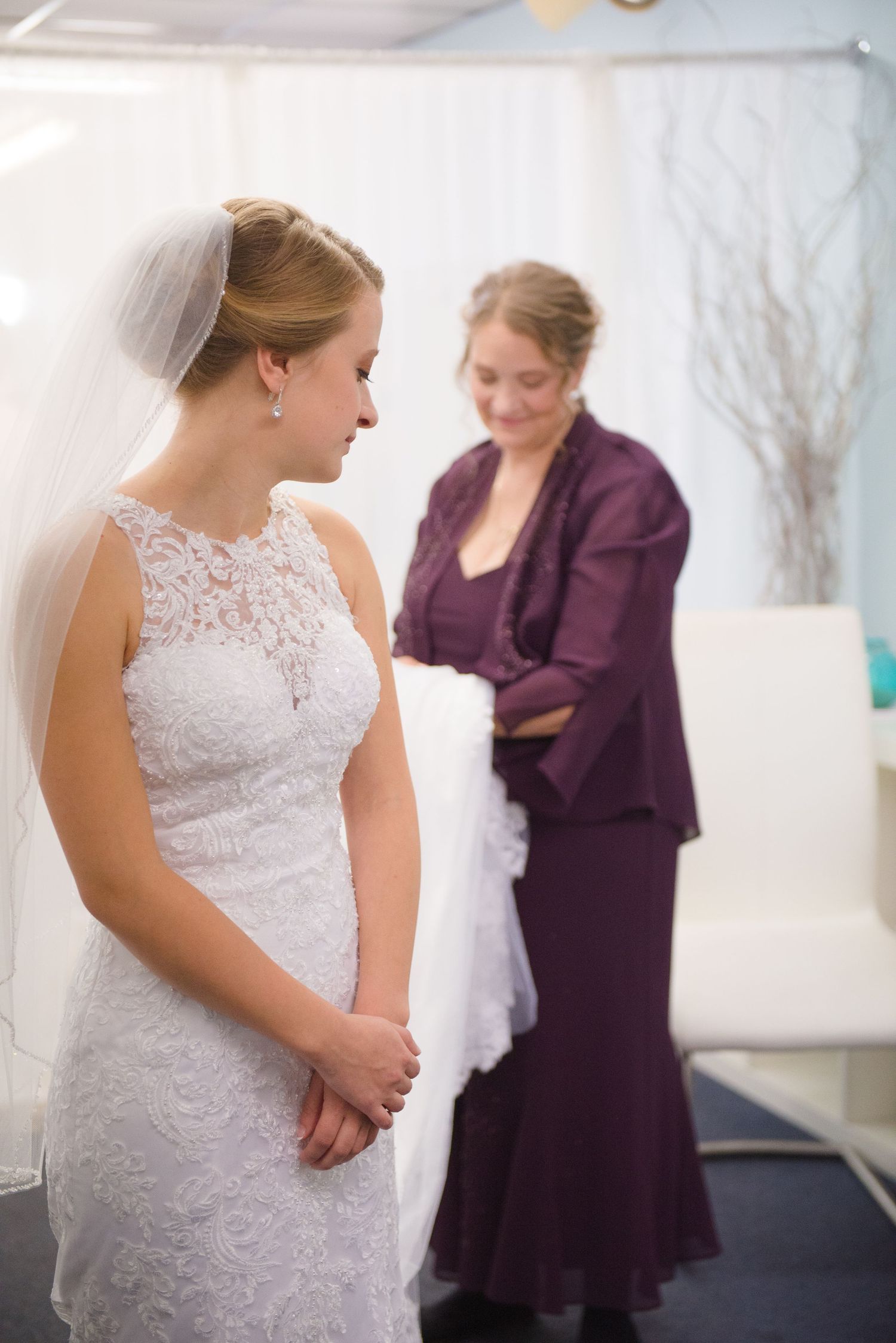 Bride in white lace gown stands while attendant in purple helps with final wedding day preparations. Andover wedding at Magestic Oaks Golf C