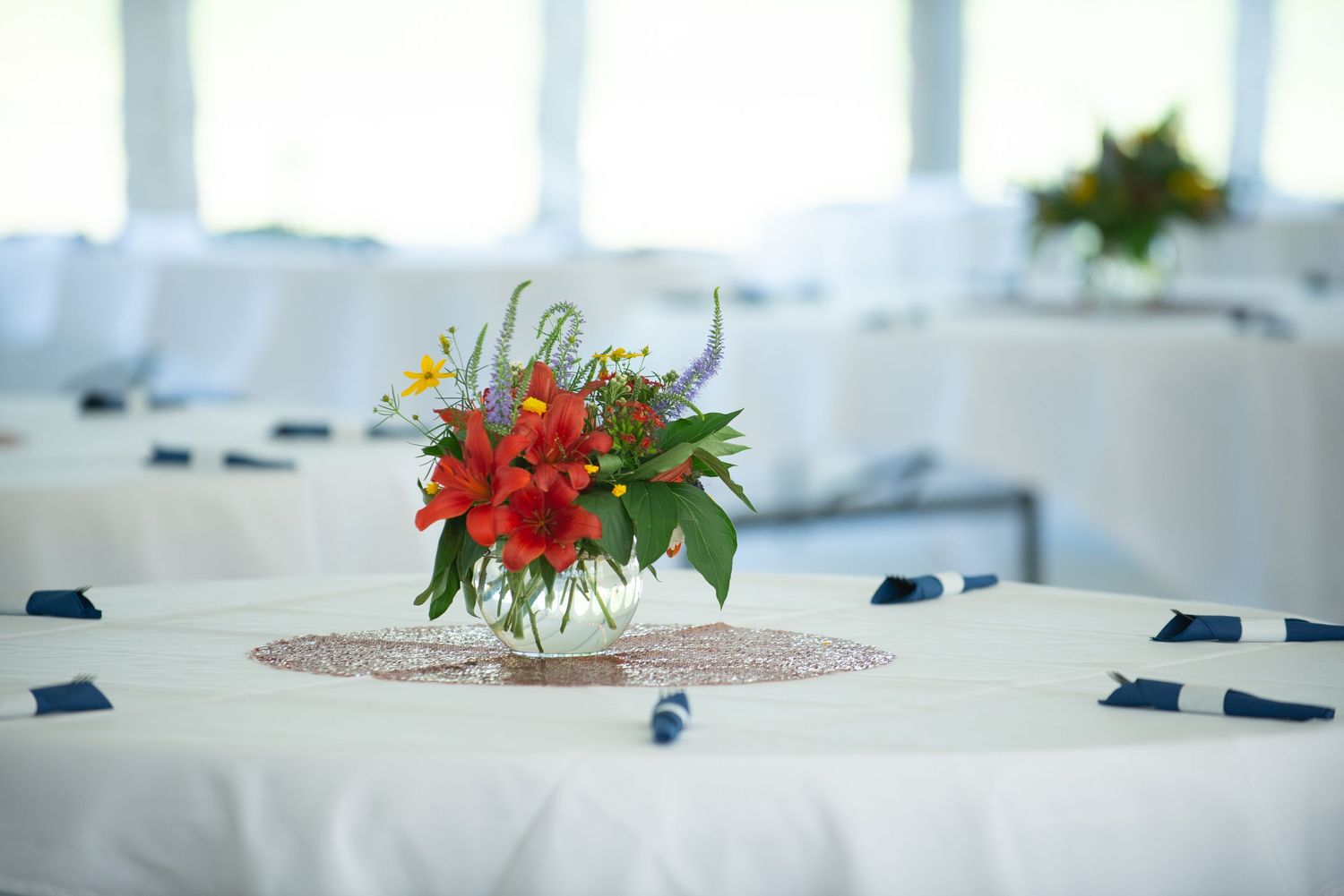 A bright red floral centerpiece with greenery sits on a white lace tablecloth in an elegant reception setting.