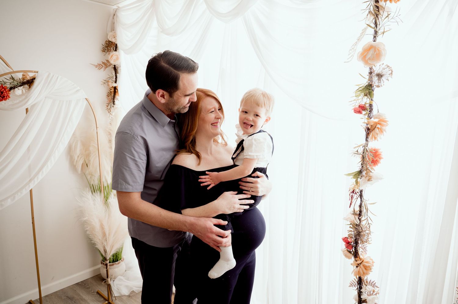 A couple stands together in front of white curtains and flowered garland while holding a baby in a gentle embrace.