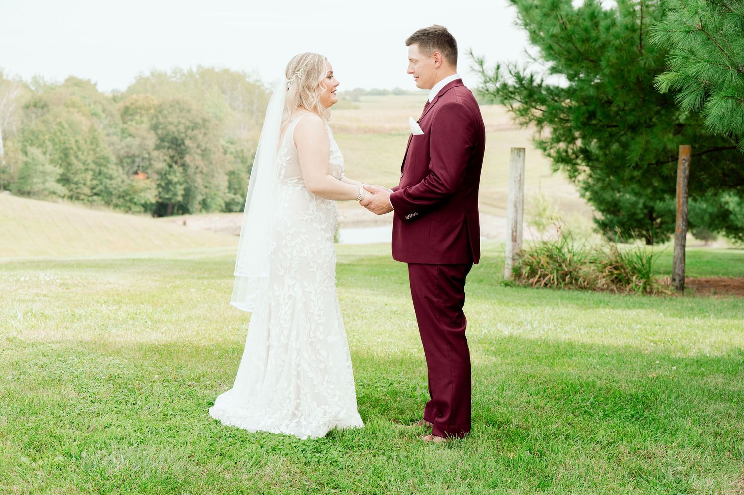 A couple in wedding attire stands together on a grassy lawn with trees in the background.