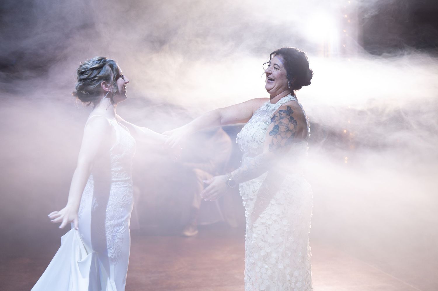 Two brides dance together in white dresses amid dramatic fog and lighting.