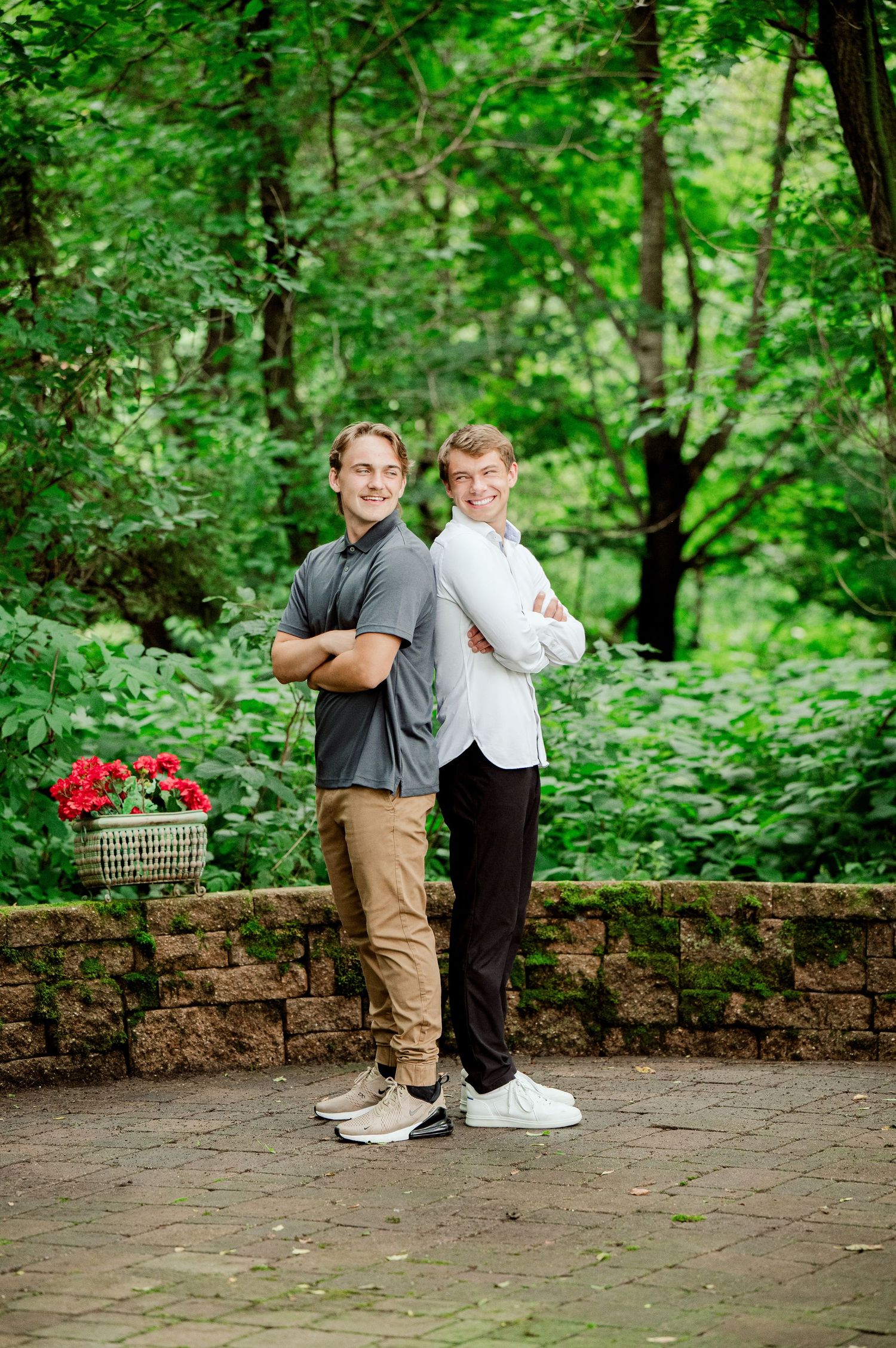 Clear Lake MN Family Photos: Two people pose back-to-back on a stone wall with a basket of red flowers in a wooded setting.