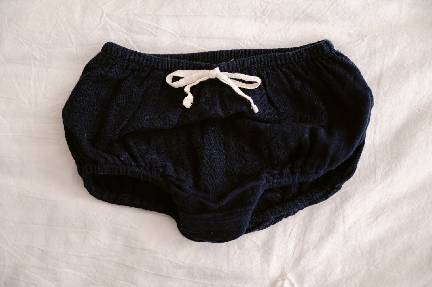 Navy blue baby bloomers with white drawstring waist lying on white fabric background.